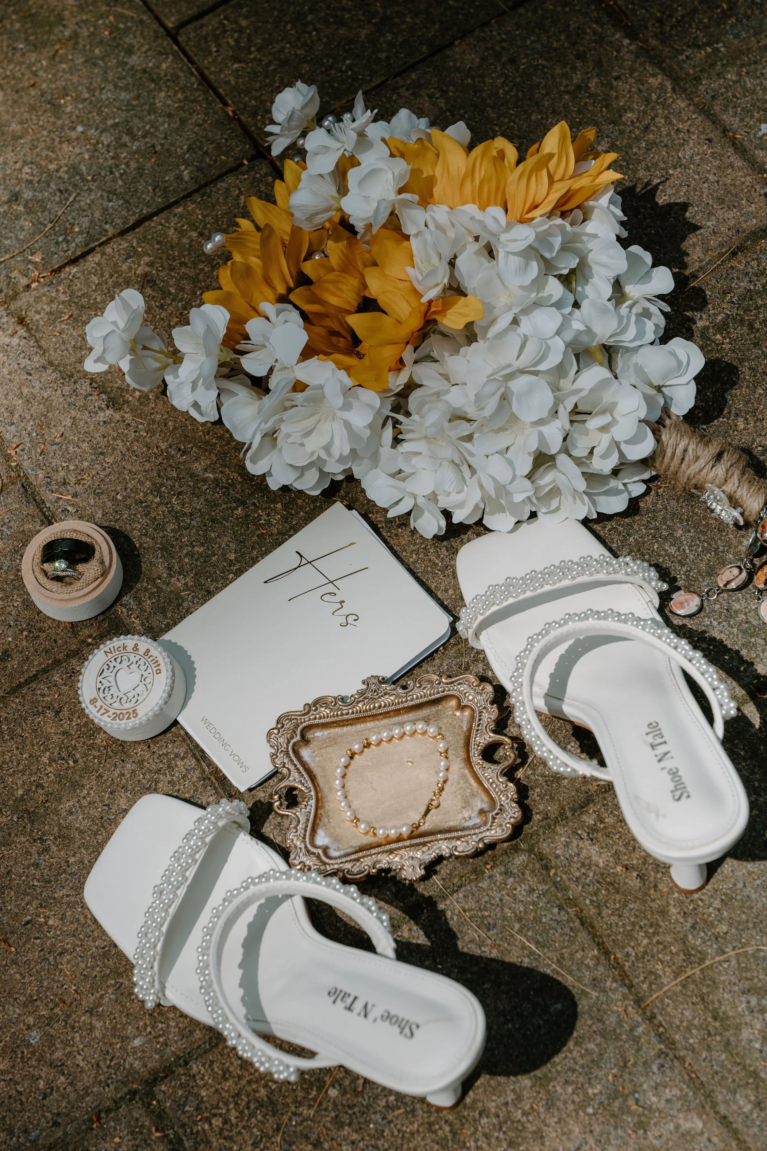A bridal arrangement with white and yellow flowers, wedding rings, pearl jewelry, white bridal shoes, and a wedding vows booklet on a paved surface for a wedding at Tunxis Country Club in Connecticut. 
