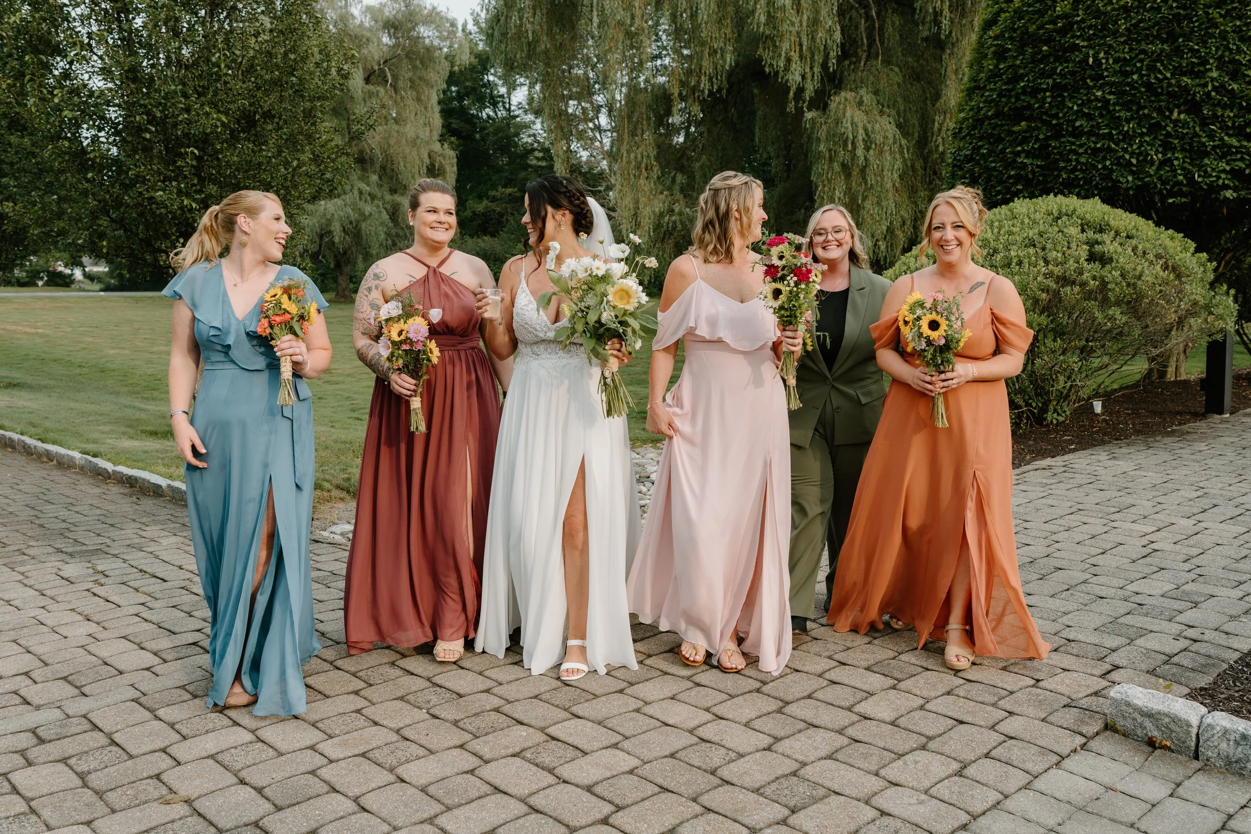 A group of seven women dressed in colorful bridesmaid and bride dresses walking outdoors on a paved path, holding bouquets, with greenery and trees in the background during a wedding at the Aqua Turf Club in Connecticut. 