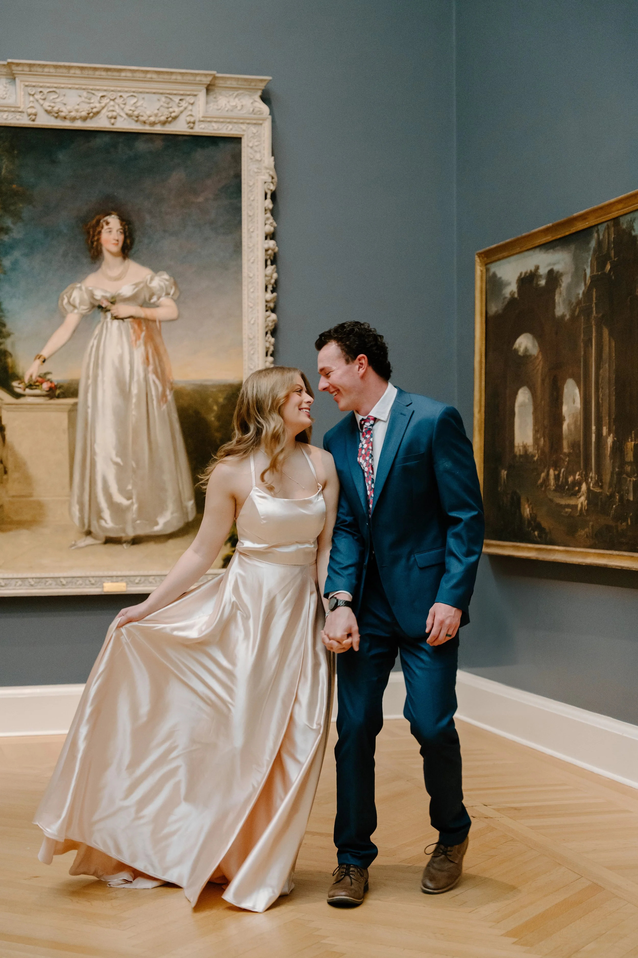A smiling couple dressed elegantly, holding hands, in an art gallery with classical paintings on the walls.