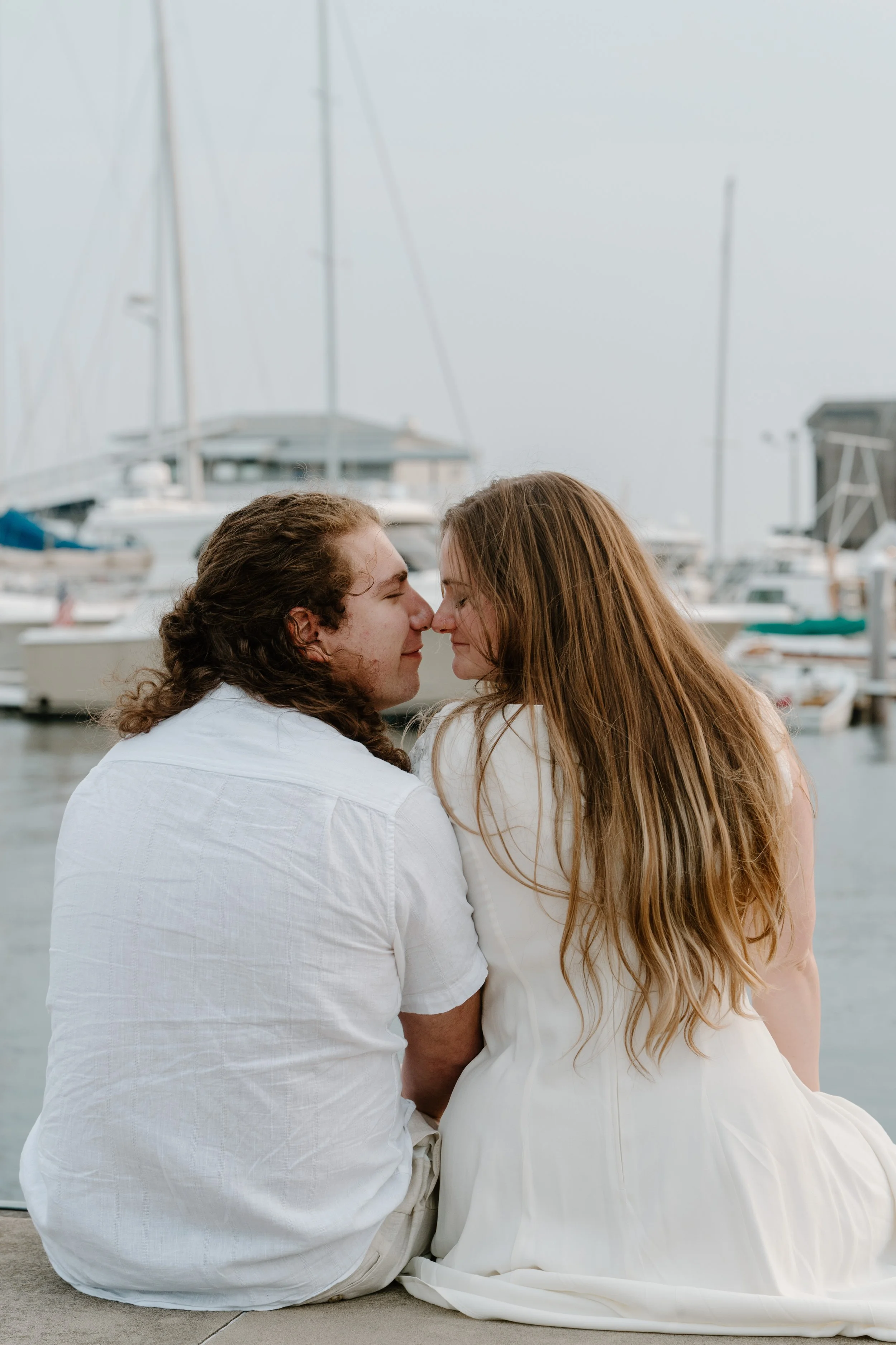 A couple sitting close together by a marina, touching foreheads with eyes closed, surrounded by boats and yachts after their elopement in Newport, Rhode Island. 