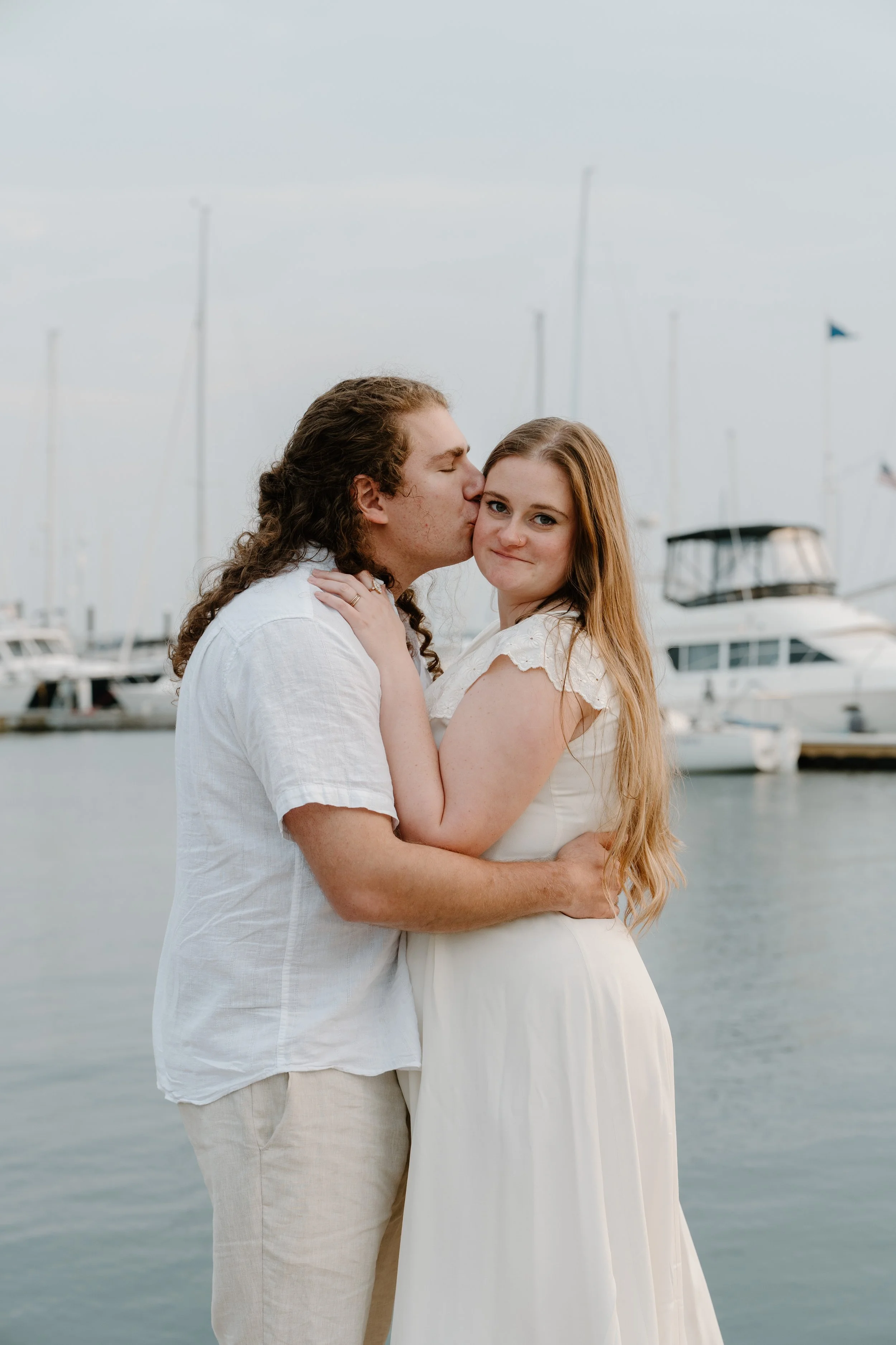 A couple embracing near a marina, with boats in the background. The man is kissing the woman's cheek, and she is smiling at the camera after an elopement at Beavertail State Park in Rhode Island. 