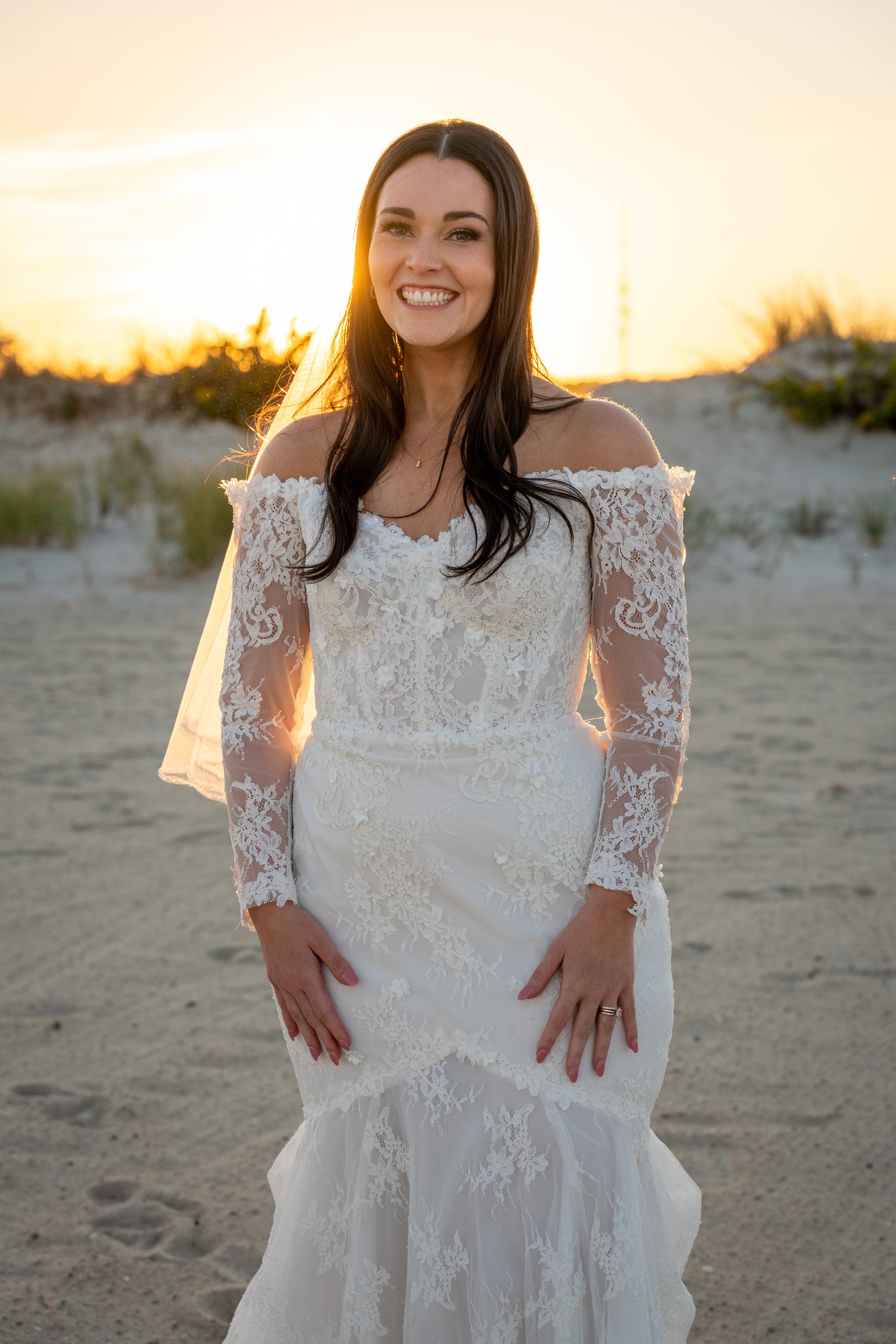 A woman in a white wedding dress standing on a sandy beach during sunset, smiling at the camera during a wedding at Red Jacket Resort in Cape Cod. 