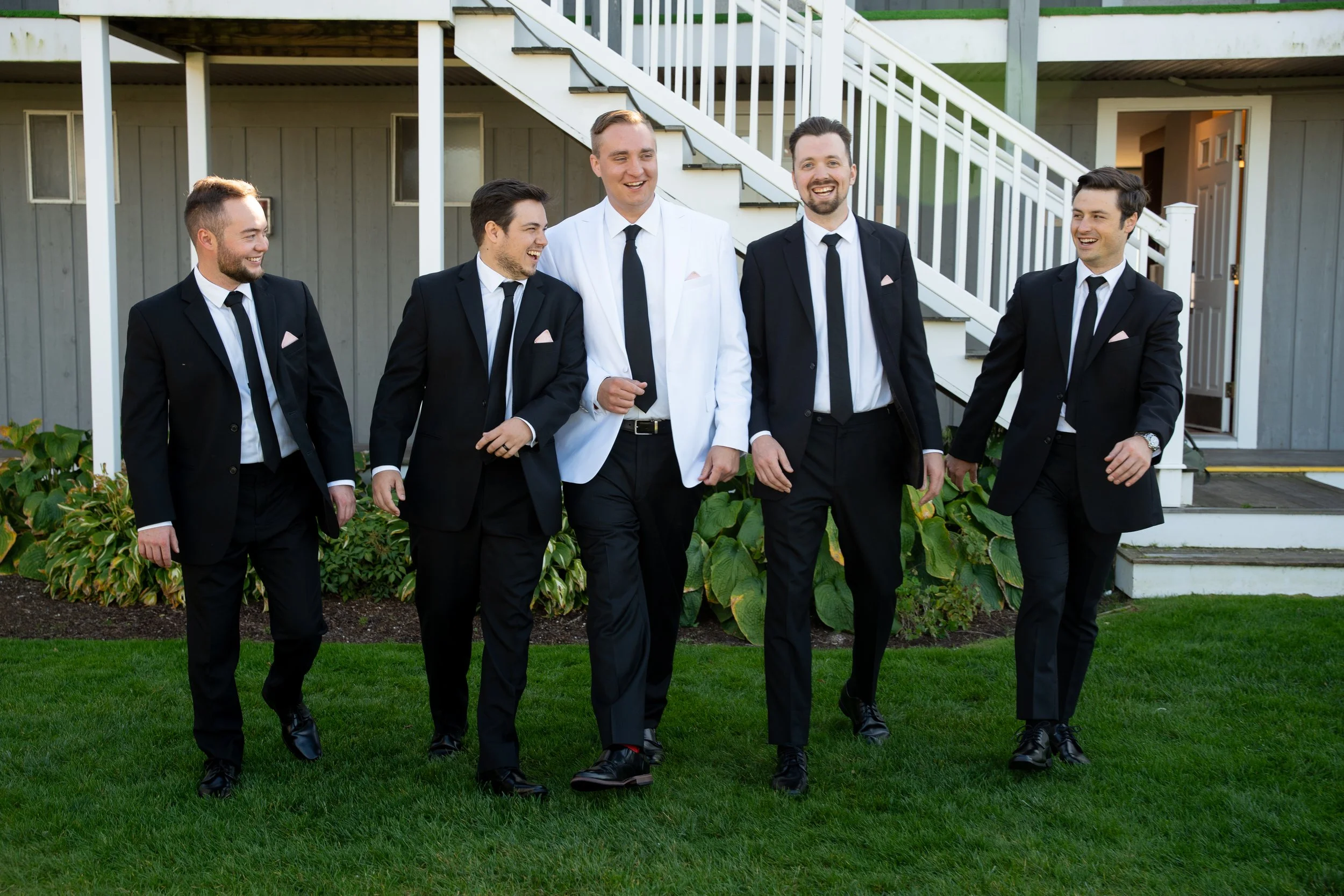 Group of five men dressed in tuxedos walking on grass outside a building with white stairs and door during a wedding at Red Jacket Resort in Cape Cod. 