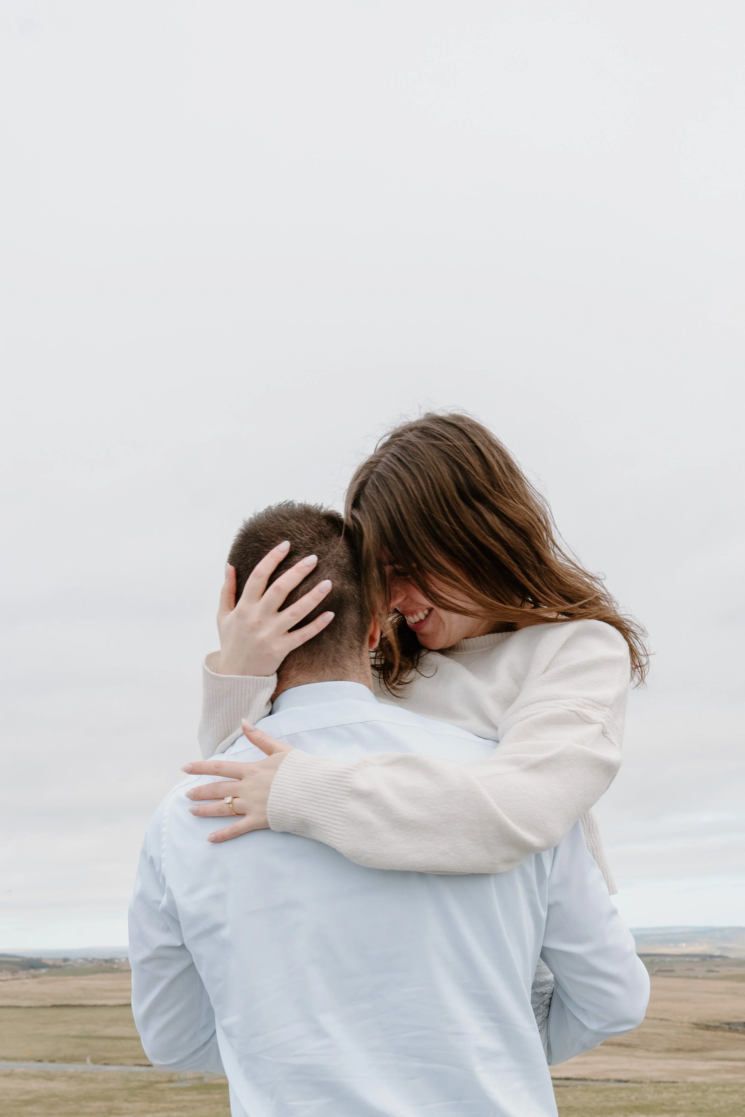 A woman and man sharing an affectionate moment outdoors, with the woman smiling and holding the man as he lifts her up. The background shows a cloudy sky and open fields for an engagement photo session in Galway, Ireland. 