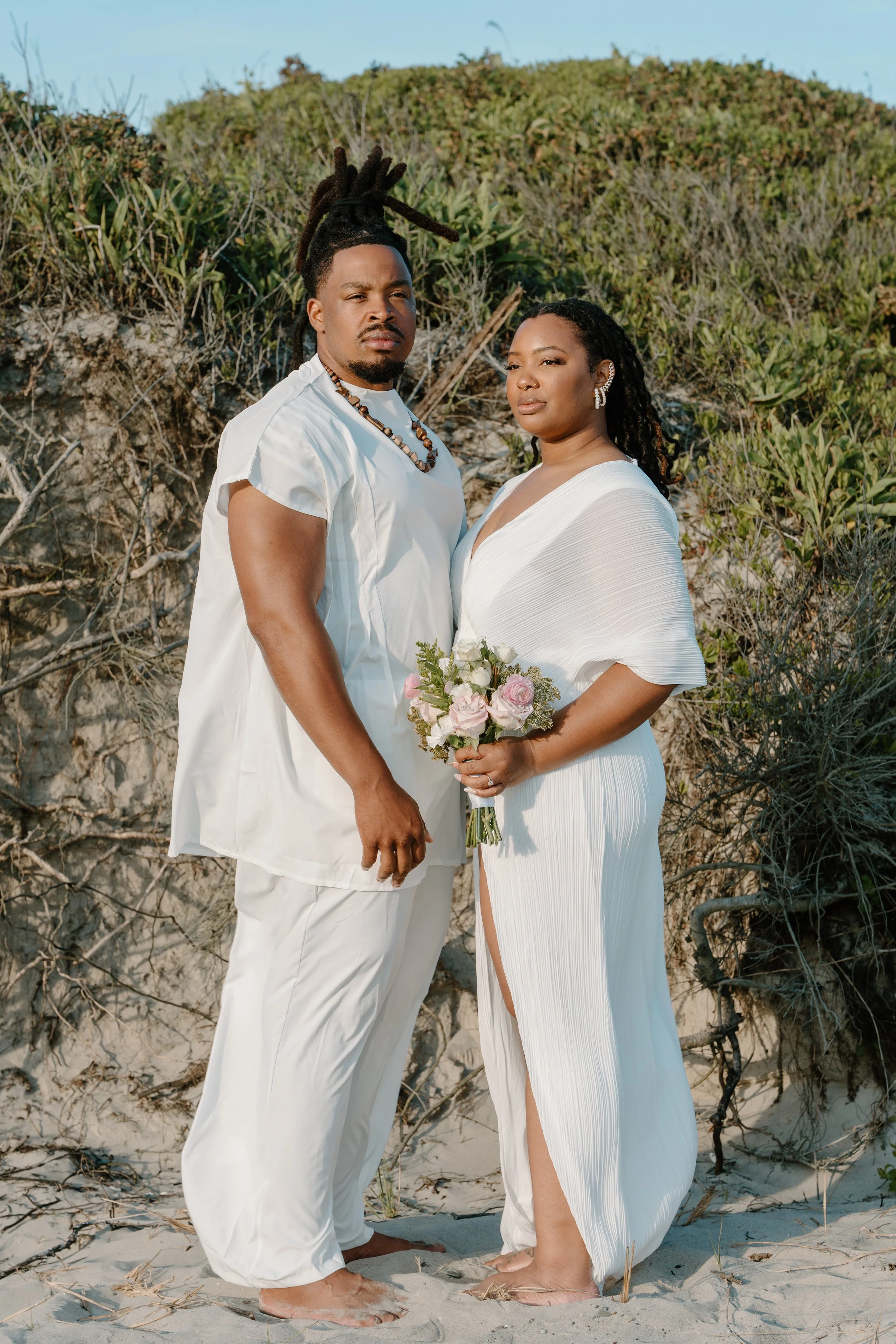 A couple dressed in white holding flowers, standing on a sandy beach with shrubs and dunes in the background at an elopement at Second Beach in Rhode Island. 