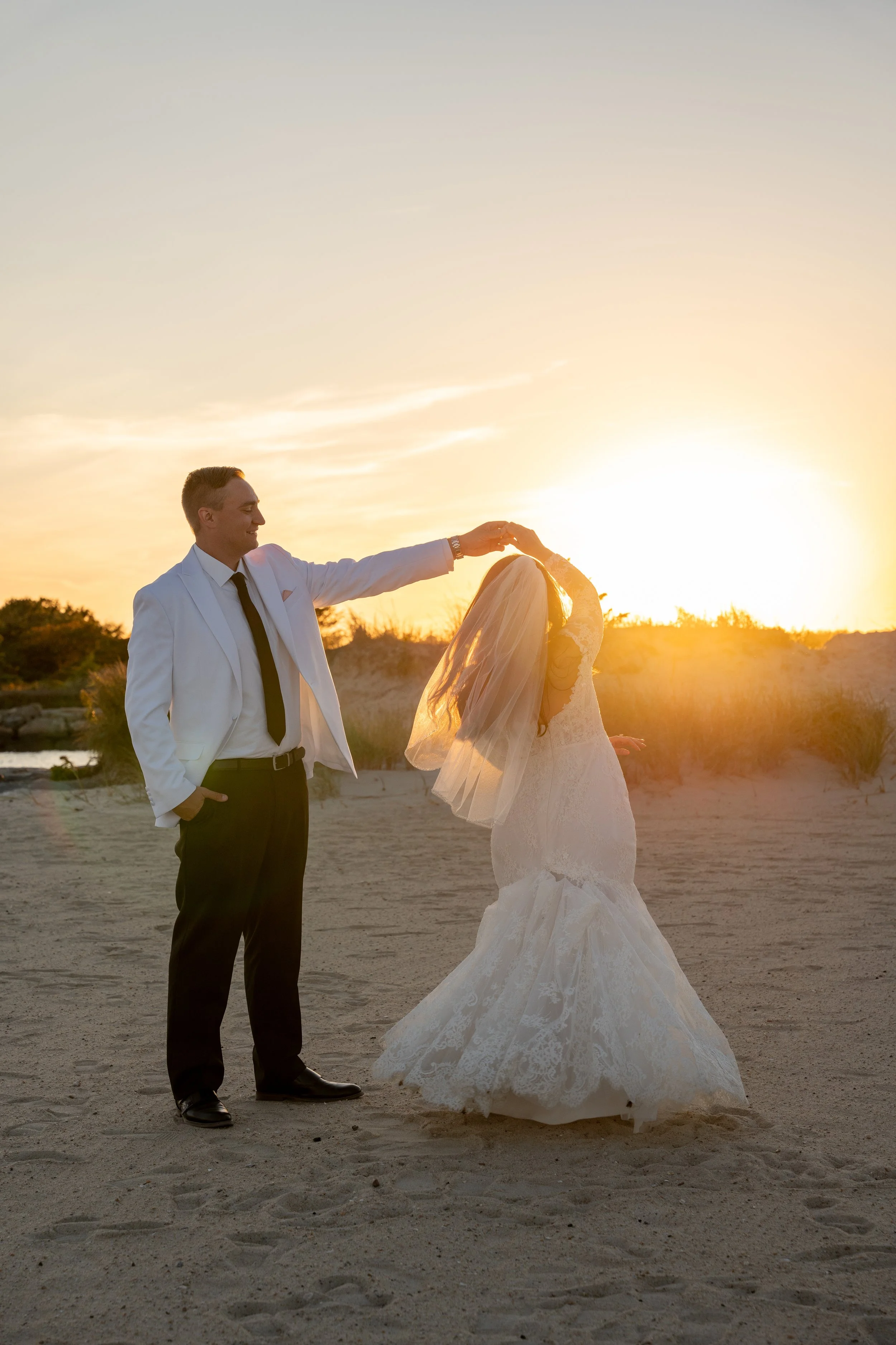 A bride and groom dance on the beach at sunset, with the groom in a white tuxedo and the bride in a lace wedding gown and veil during a wedding at Red Jacket Resort in Cape Cod. 