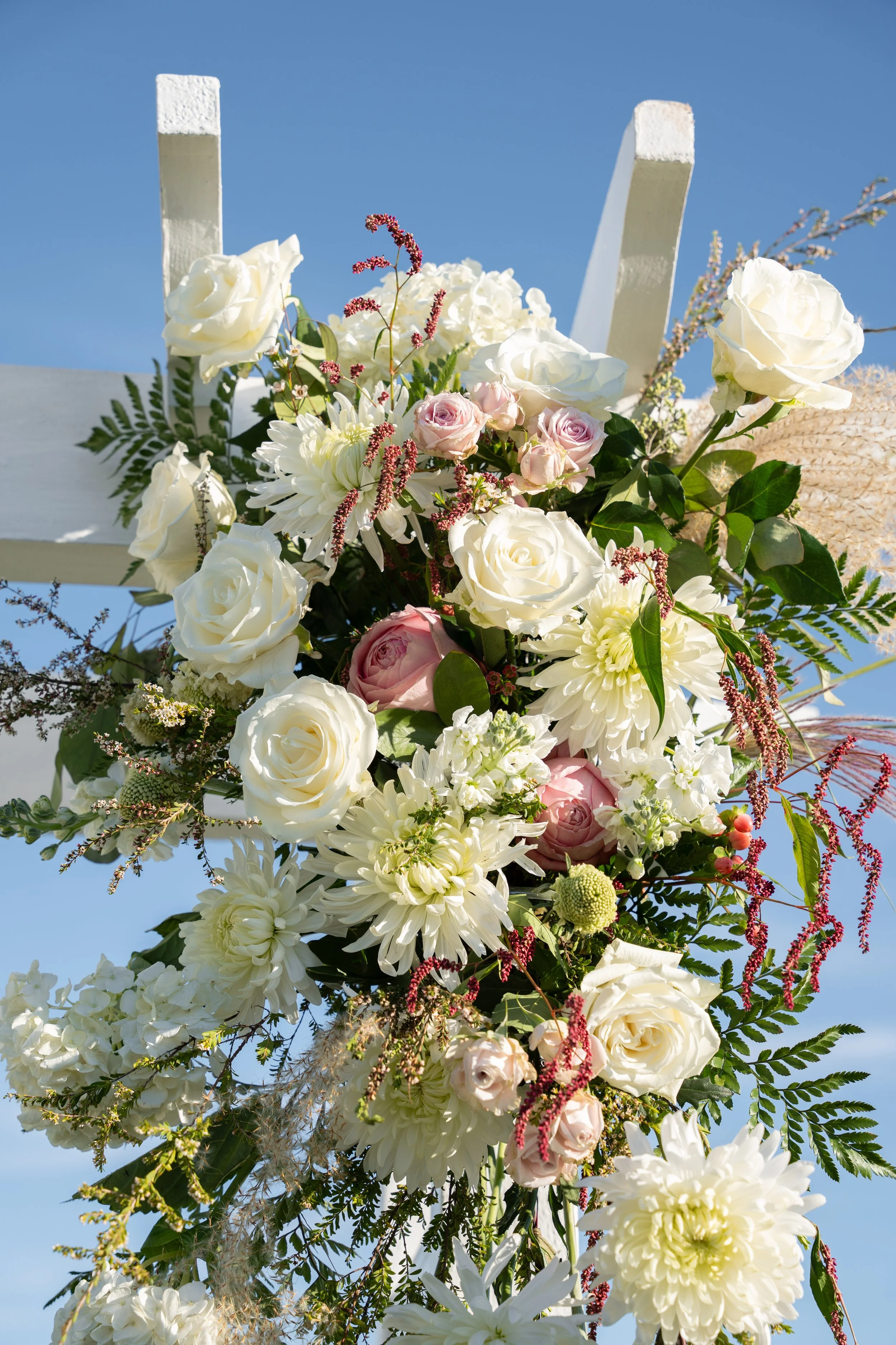 A vertical floral arrangement featuring white roses, pale pink roses, white dahlias, and various greenery against a blue sky during a wedding at Red Jacket Resort in Cape Cod. 