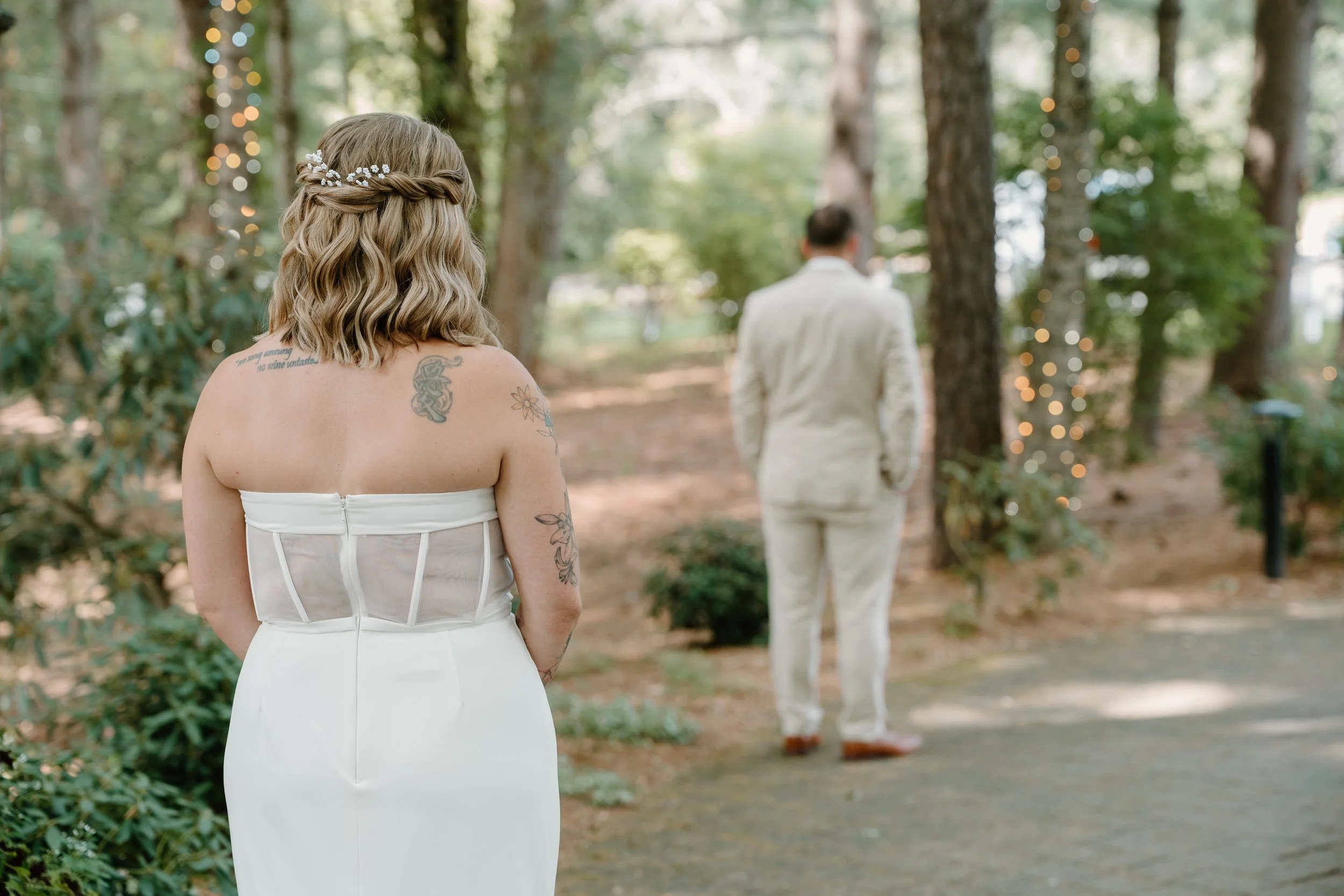 A woman in a white strapless dress with tattoos on her shoulders and arms, standing outdoors in a wooded area with bokeh string lights, watching a man in a light-colored suit with his back turned for a wedding at Tunxis Country Club in Connecticut. 