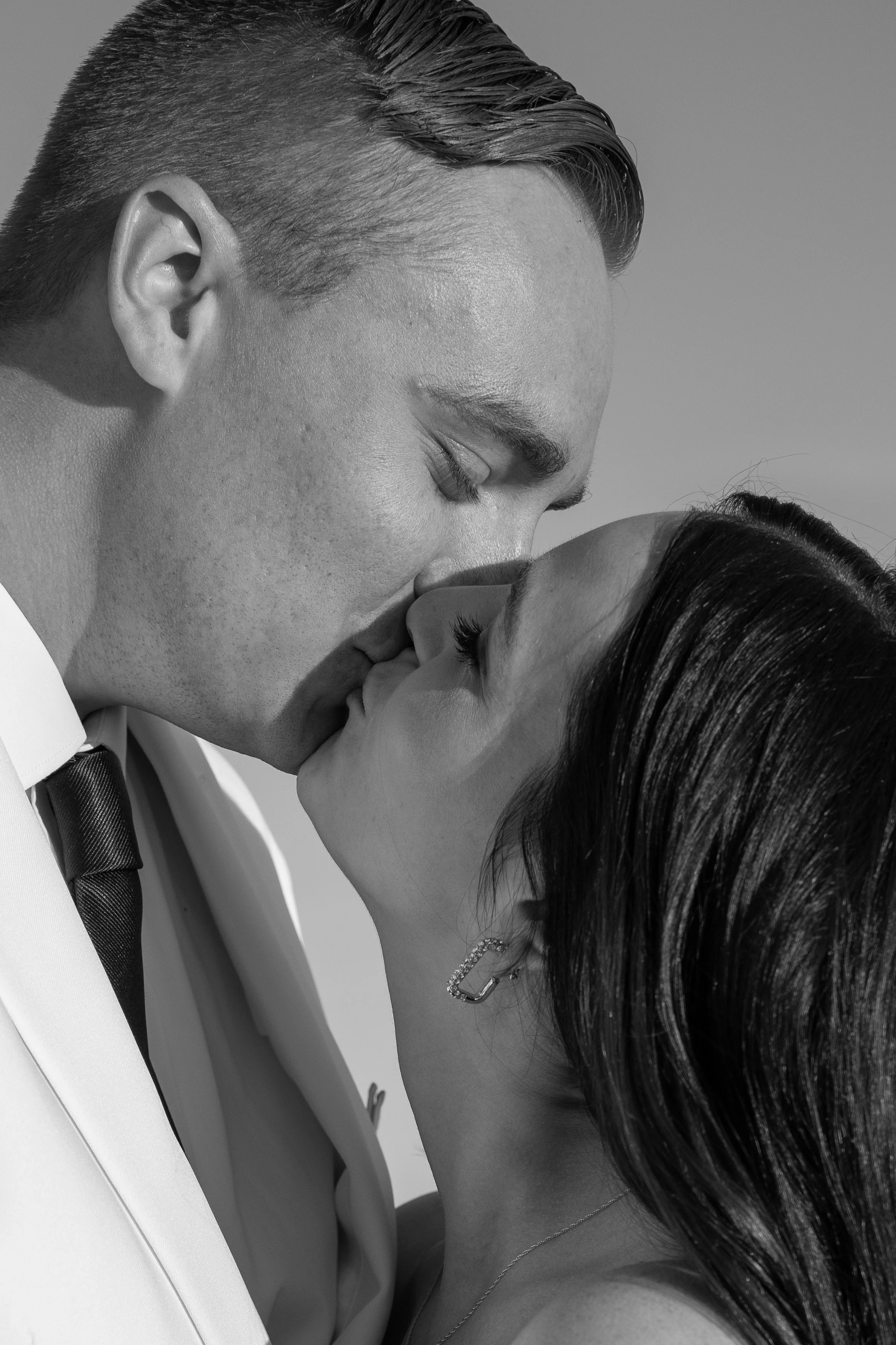 A black and white photo of a couple sharing a kiss, with the man wearing a suit and tie and the woman wearing earrings and a necklace during a wedding at Red Jacket Resort in Cape Cod. 