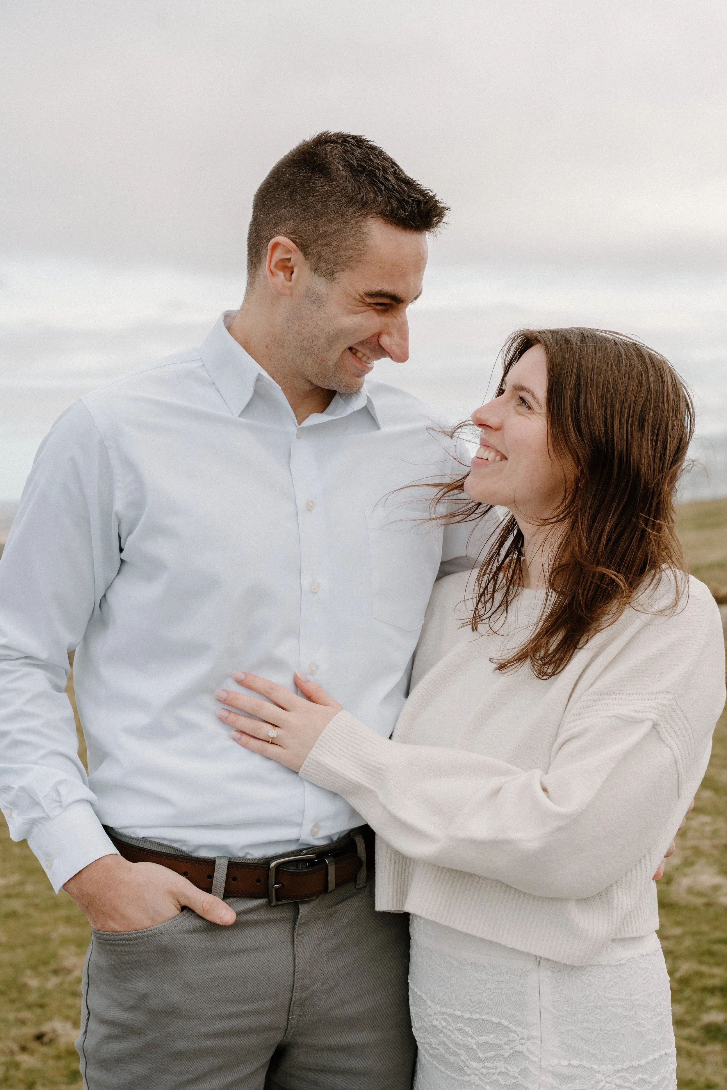 A happy couple smiling and looking at each other outdoors on a cloudy day, embracing each other for an engagement photo session in Galway, Ireland. 