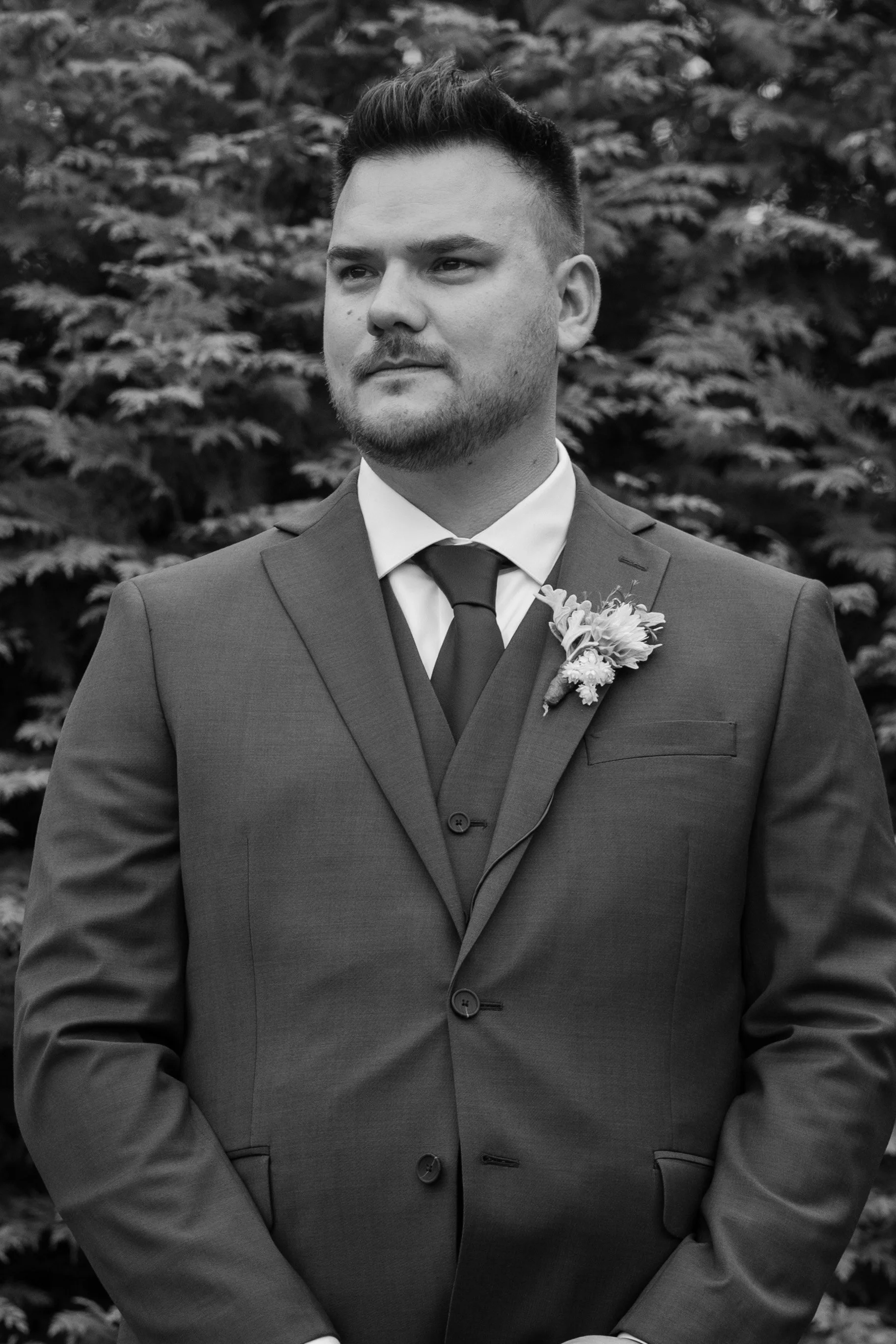 Black and white photo of a man in a suit with a boutonniere, standing outdoors in front of foliage during a wedding at the Aqua Turf Club in Connecticut. 