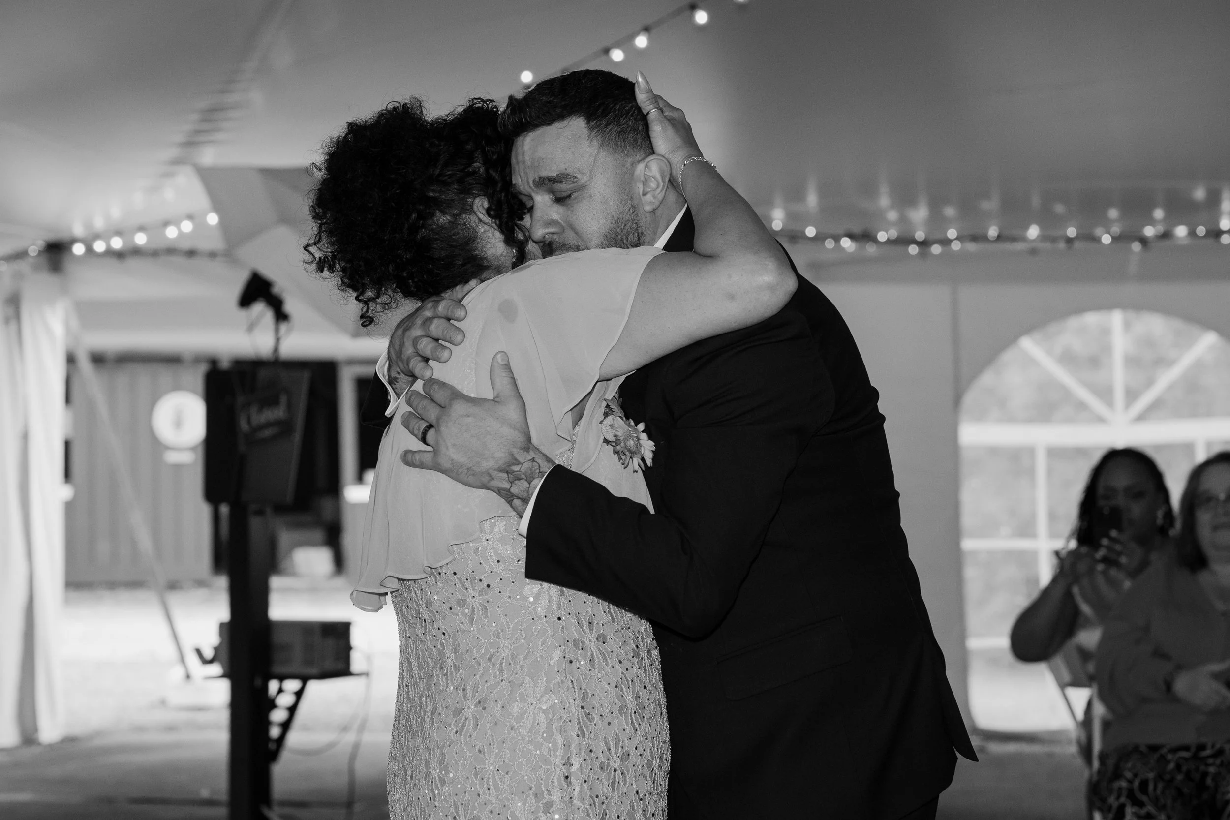 A mother and son share an emotional hug during a wedding reception in a tent, with guests watching and string lights overhead for a wedding at Holiday Hill Day Camp in Connecticut.