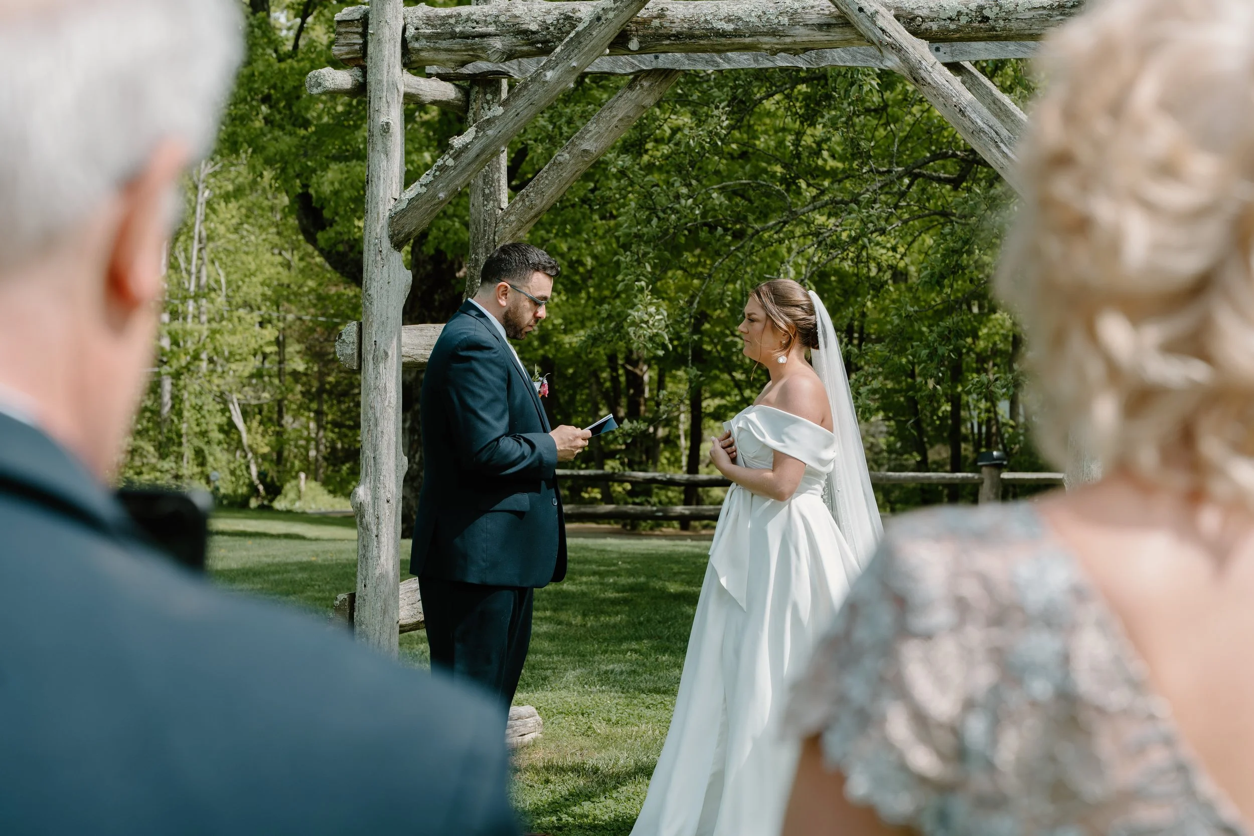 A wedding ceremony outdoors with the bride and groom standing under a rustic wooden arch. The groom is reading vows from a card, while the bride listens with her hands clasped for a wedding at Holiday Hill Day Camp in Connecticut.