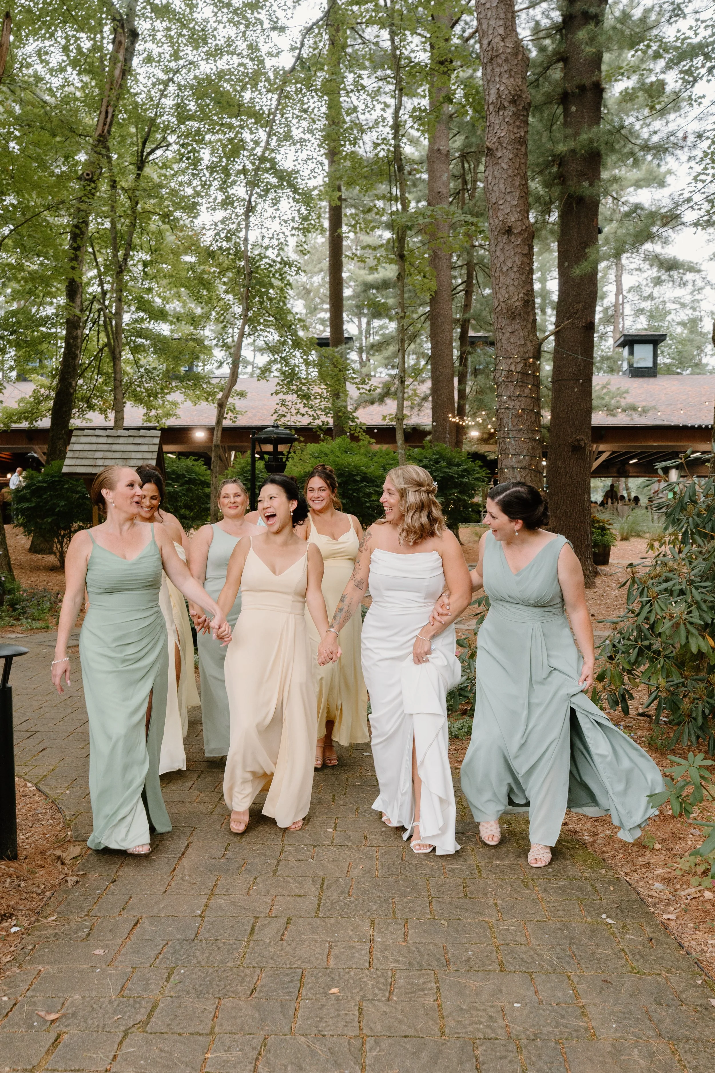 Group of women in pastel dresses walking and laughing outdoors in a wooded area during daytime for a wedding at Tunxis Country Club in Connecticut. 