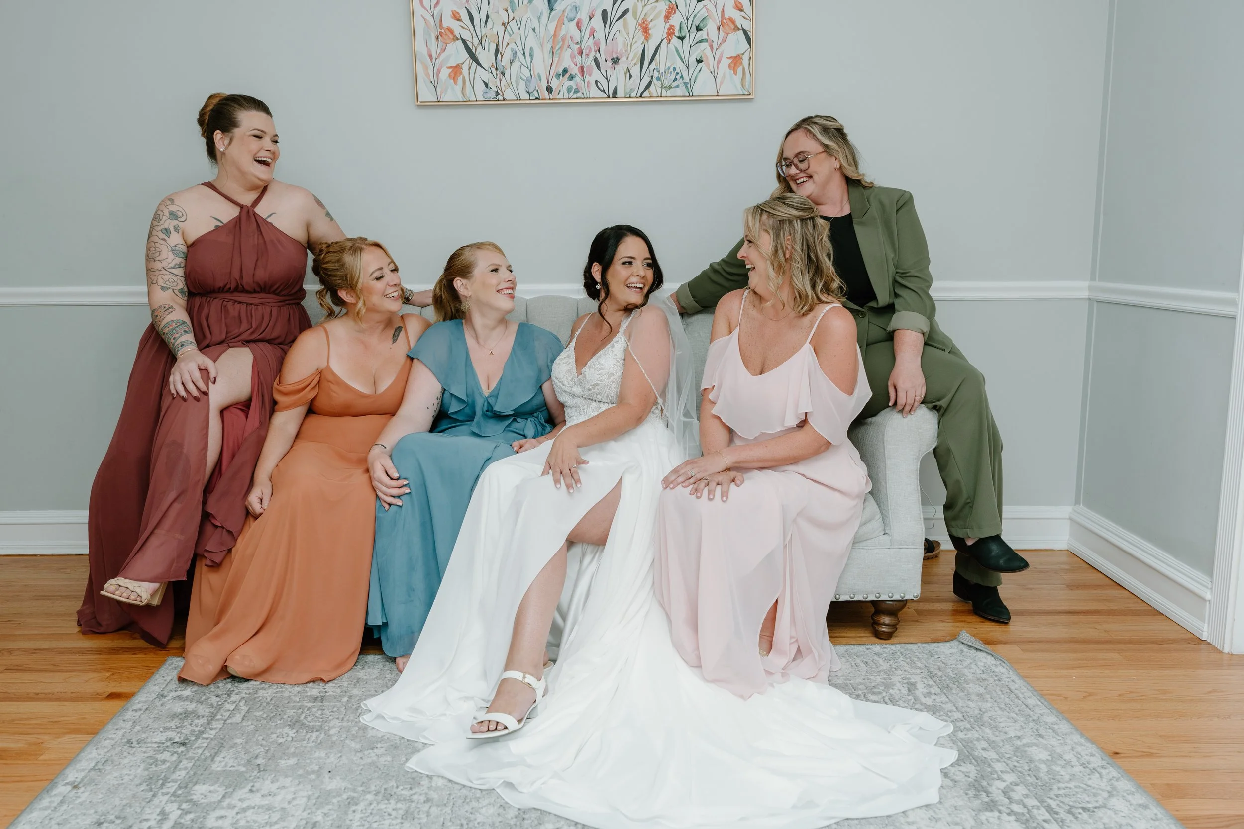 Group of women, including a bride in a white wedding gown, sitting and standing together in a living room, smiling and laughing during a wedding at the Aqua Turf Club in Connecticut. 