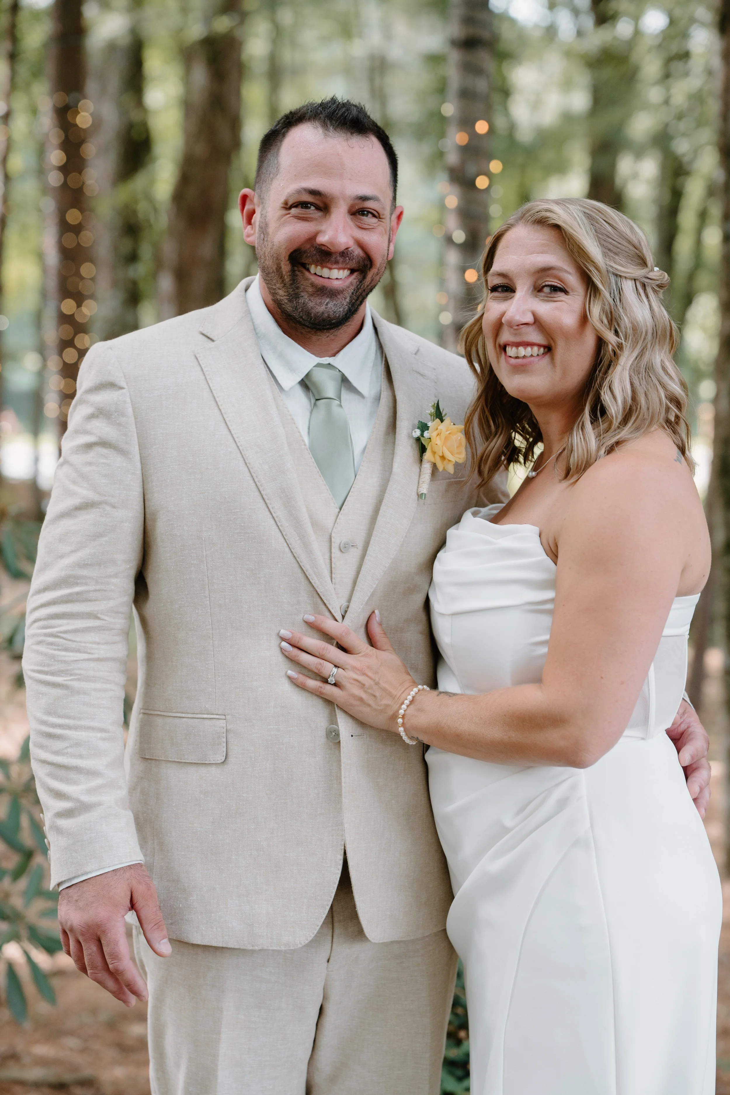 A smiling couple dressed in wedding attire, standing outdoors in a wooded area with string lights in the background for a wedding at Tunxis Country Club in Connecticut. 