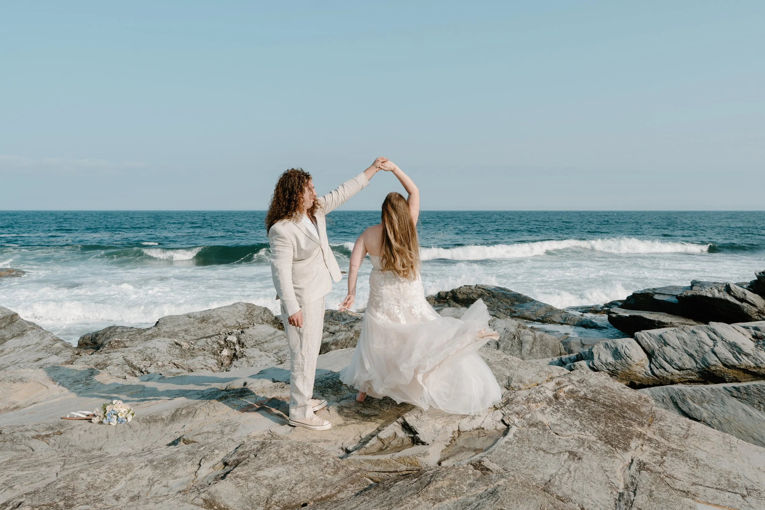 A couple dancing on rocky beach, woman in wedding dress, man in light-colored suit, ocean waves in background at an elopement at Beavertail State Park in Rhode Island. 