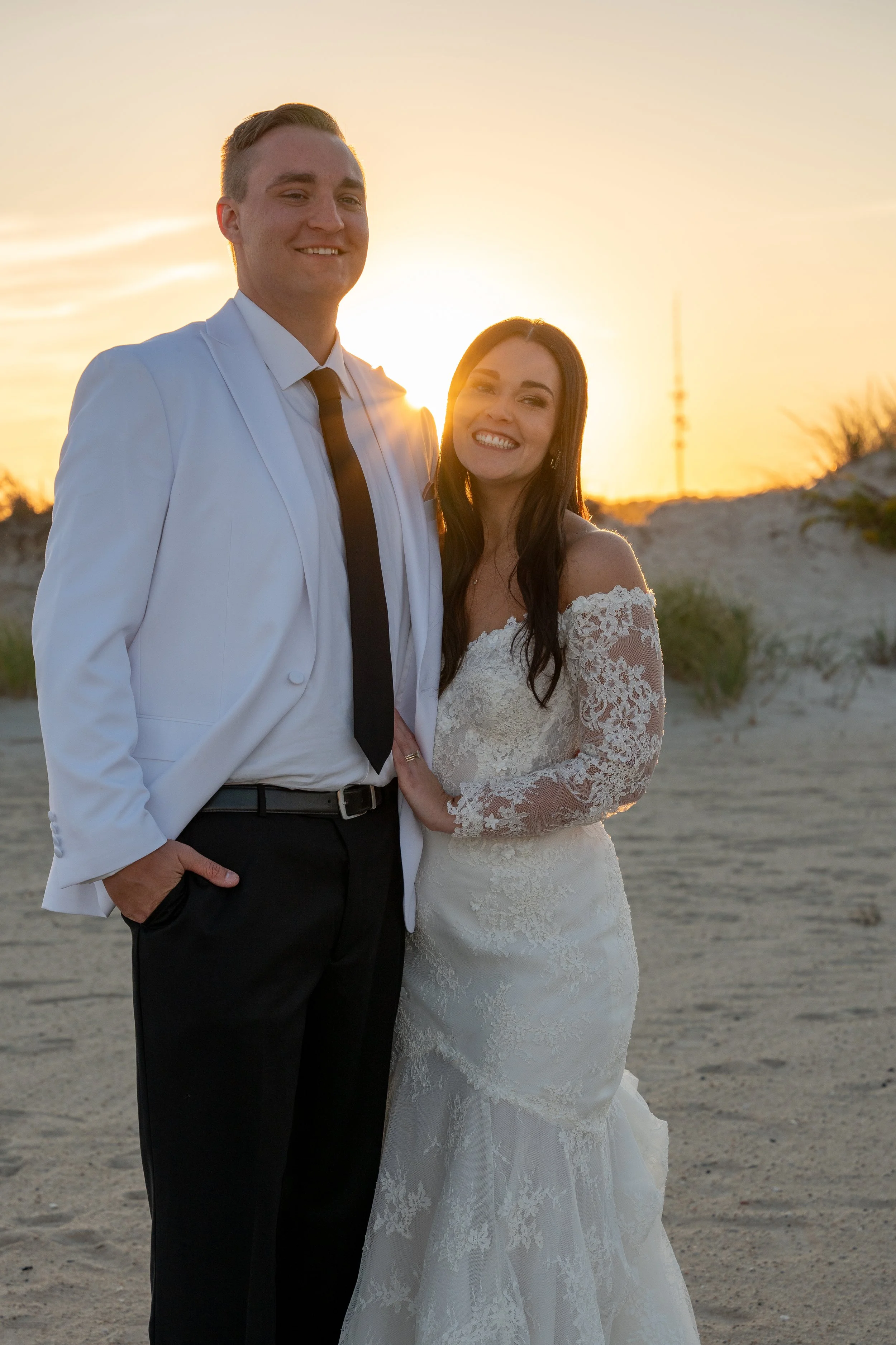 A smiling couple in wedding attire standing on a sandy area at sunset, with ocean dunes and a communication tower in the background during a wedding at Red Jacket Resort in Cape Cod. 