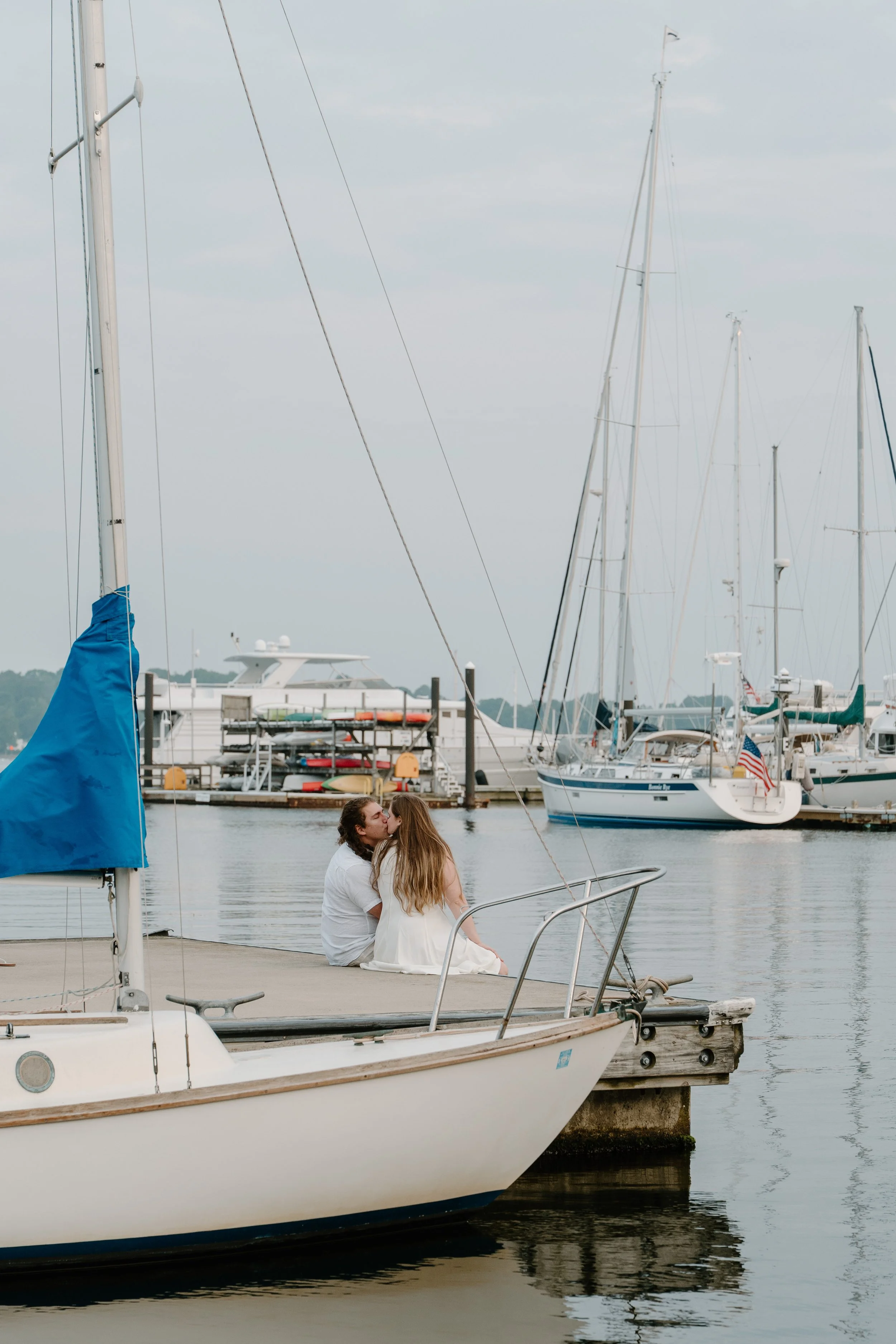 Couple sitting on a dock kissing with sailboats and yachts in the background at a marina after an elopement at Beavertail State Park in Rhode Island. 