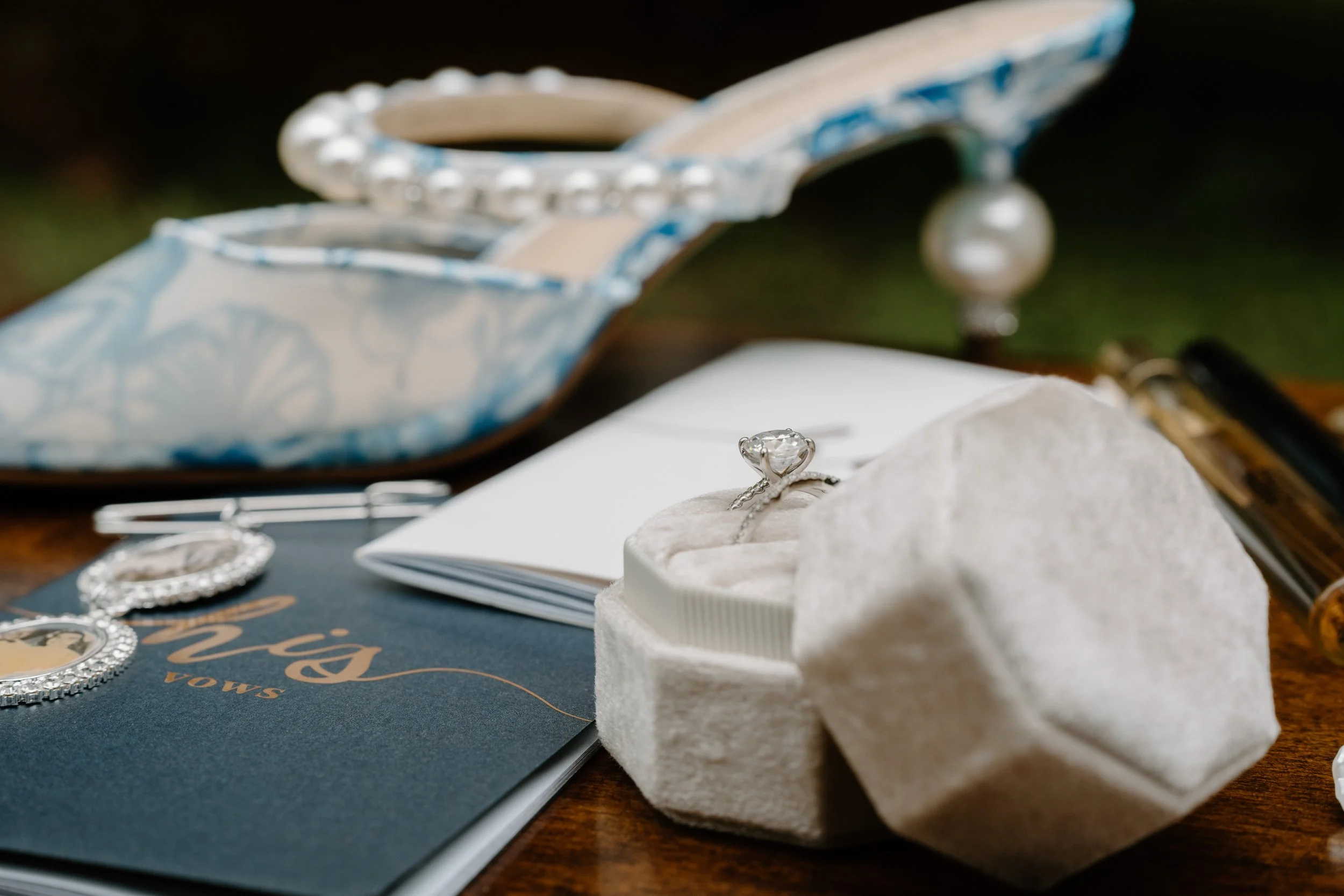 Close-up of a diamond engagement ring in a plush white ring box on a table surrounded by blue shoes with pearl accents, jewelry, and a dark blue guestbook with gold lettering for a wedding at Holiday Hill Day Camp in Connecticut.