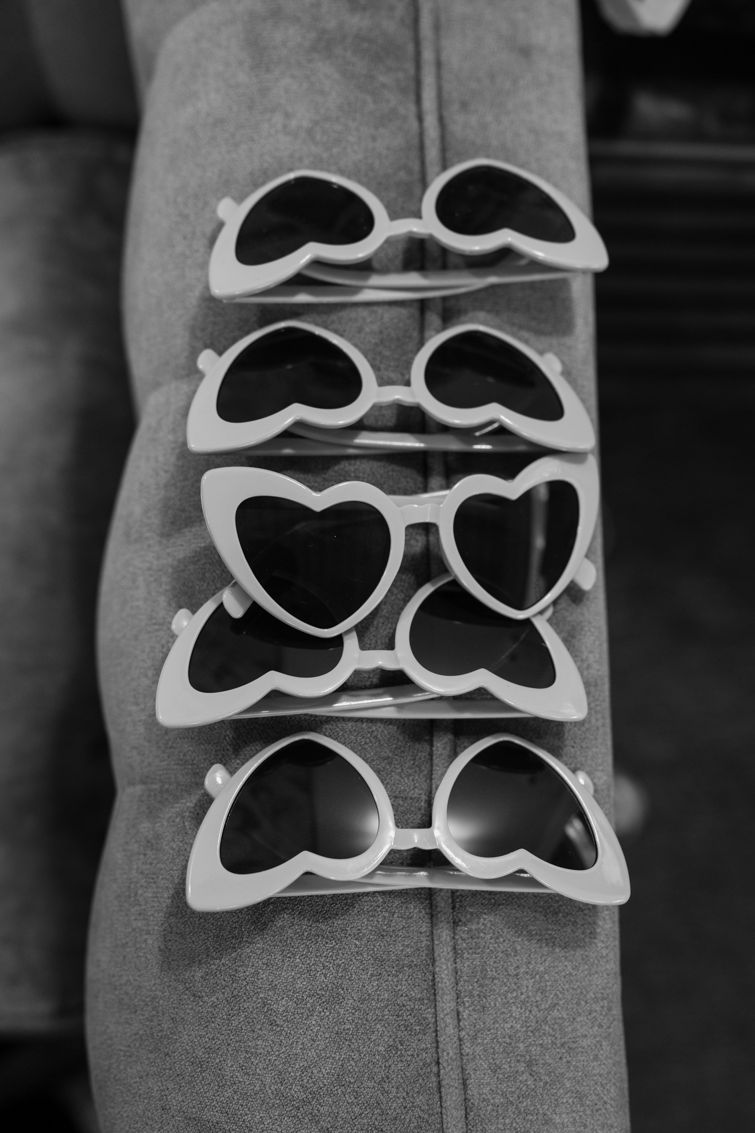 Four pairs of white heart-shaped sunglasses with dark lenses stacked on a gray cushioned surface for a wedding at Tunxis Country Club in Connecticut. 