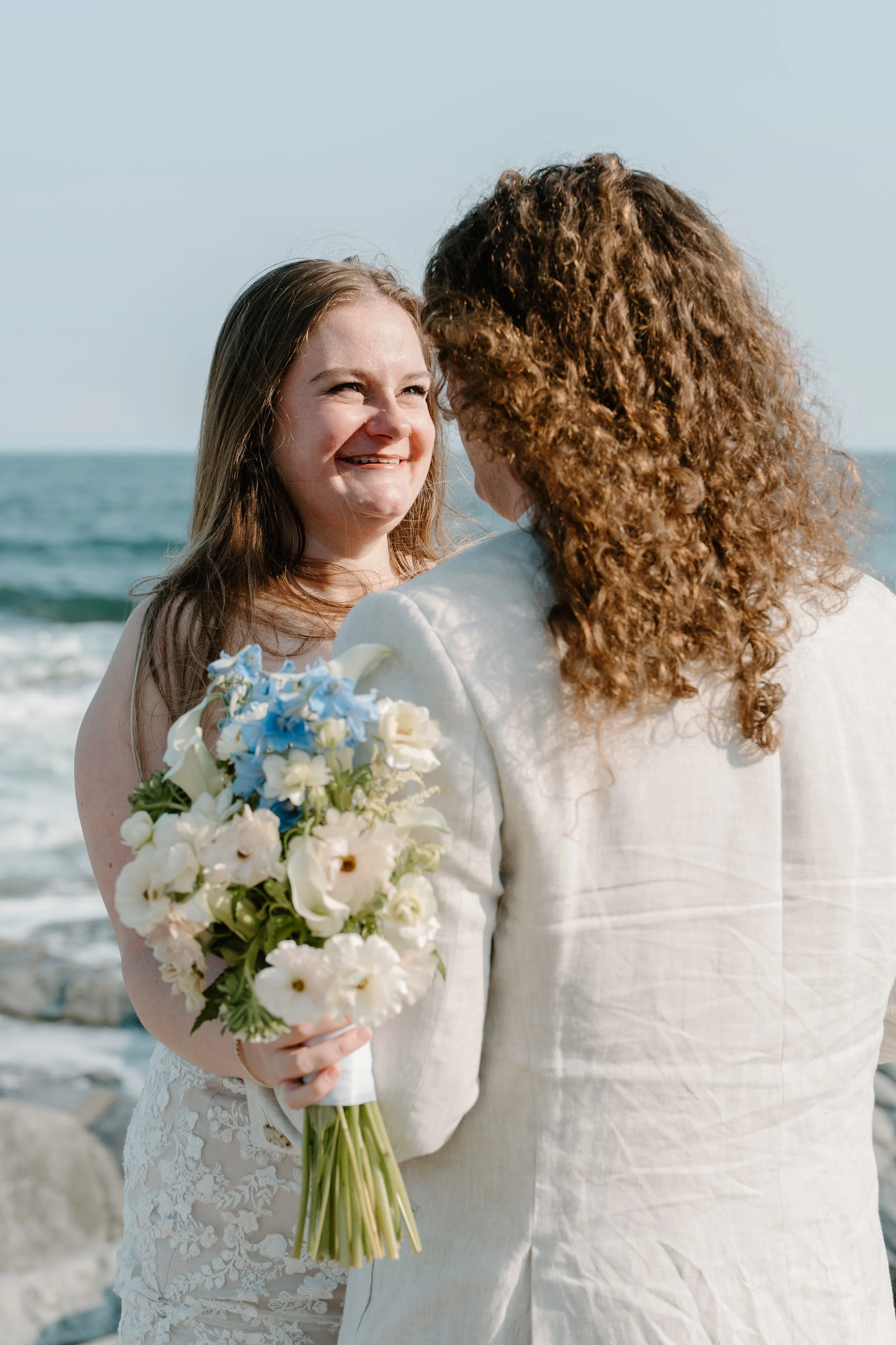 A woman with long hair holding a bouquet of white and blue flowers, facing another woman with curly hair, on a beach at an elopement at Beavertail State Park in Rhode Island. 