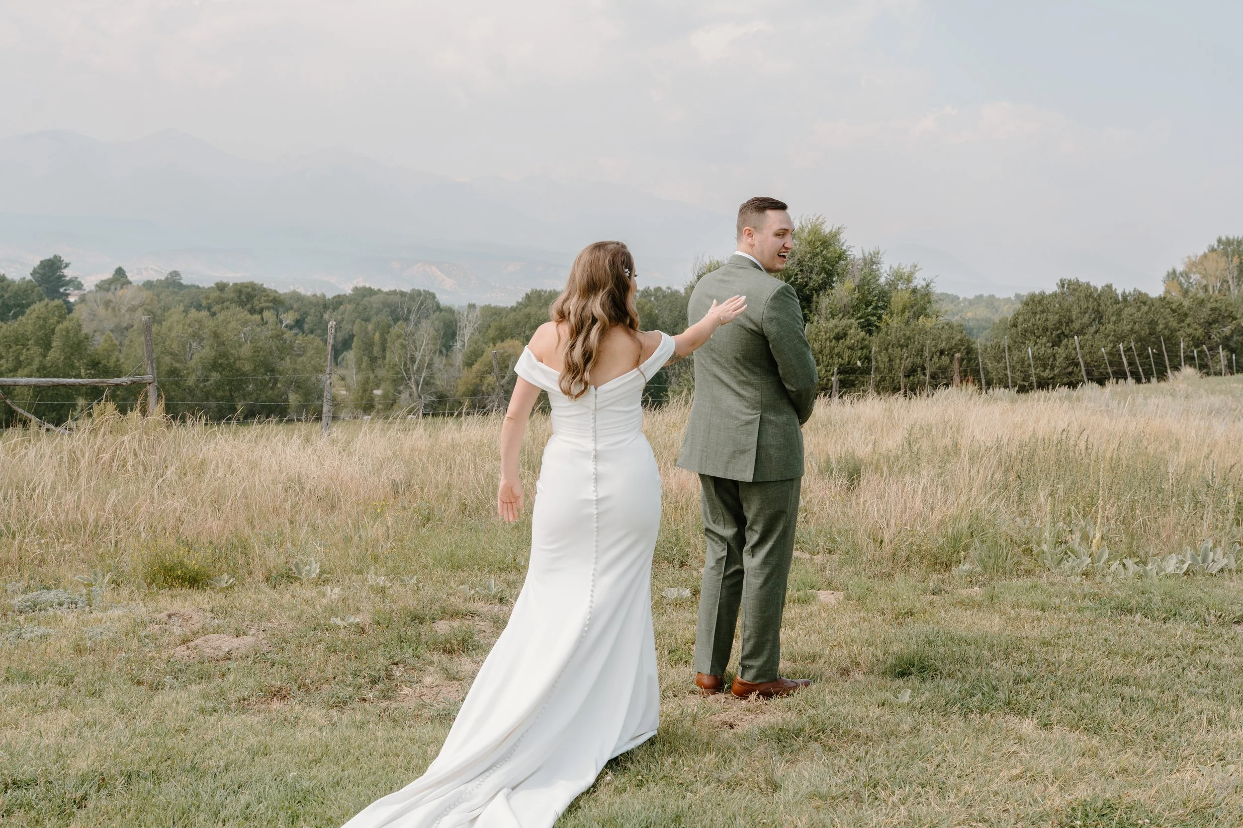 A bride in a white wedding dress and a groom in a gray suit walking through a grassy field with trees and mountains in the background at a wedding at Everett Ranch in Salida, Colorado.