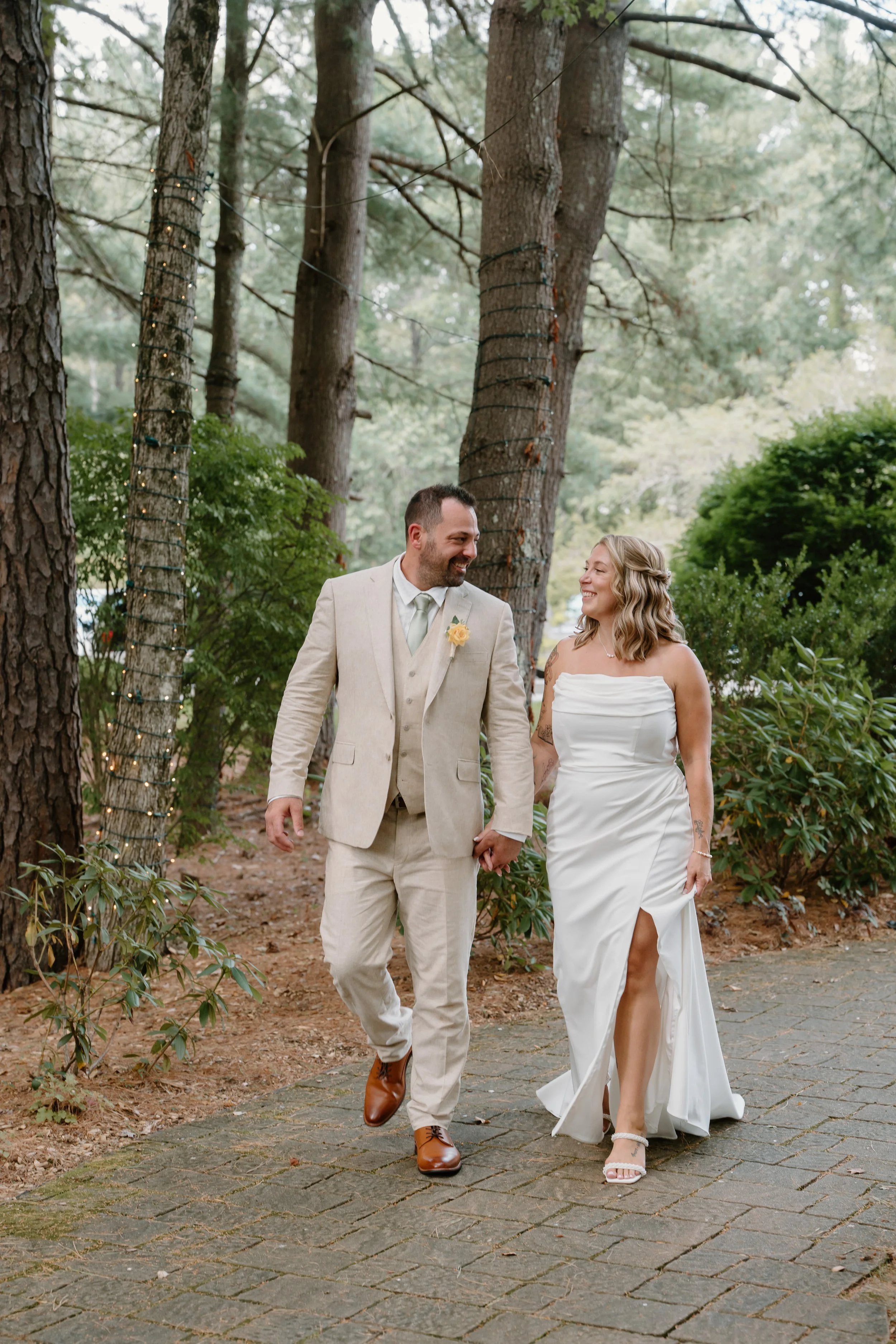 A couple in wedding attire walking hand in hand outdoors surrounded by tall trees and greenery for a wedding at Tunxis Country Club in Connecticut. 