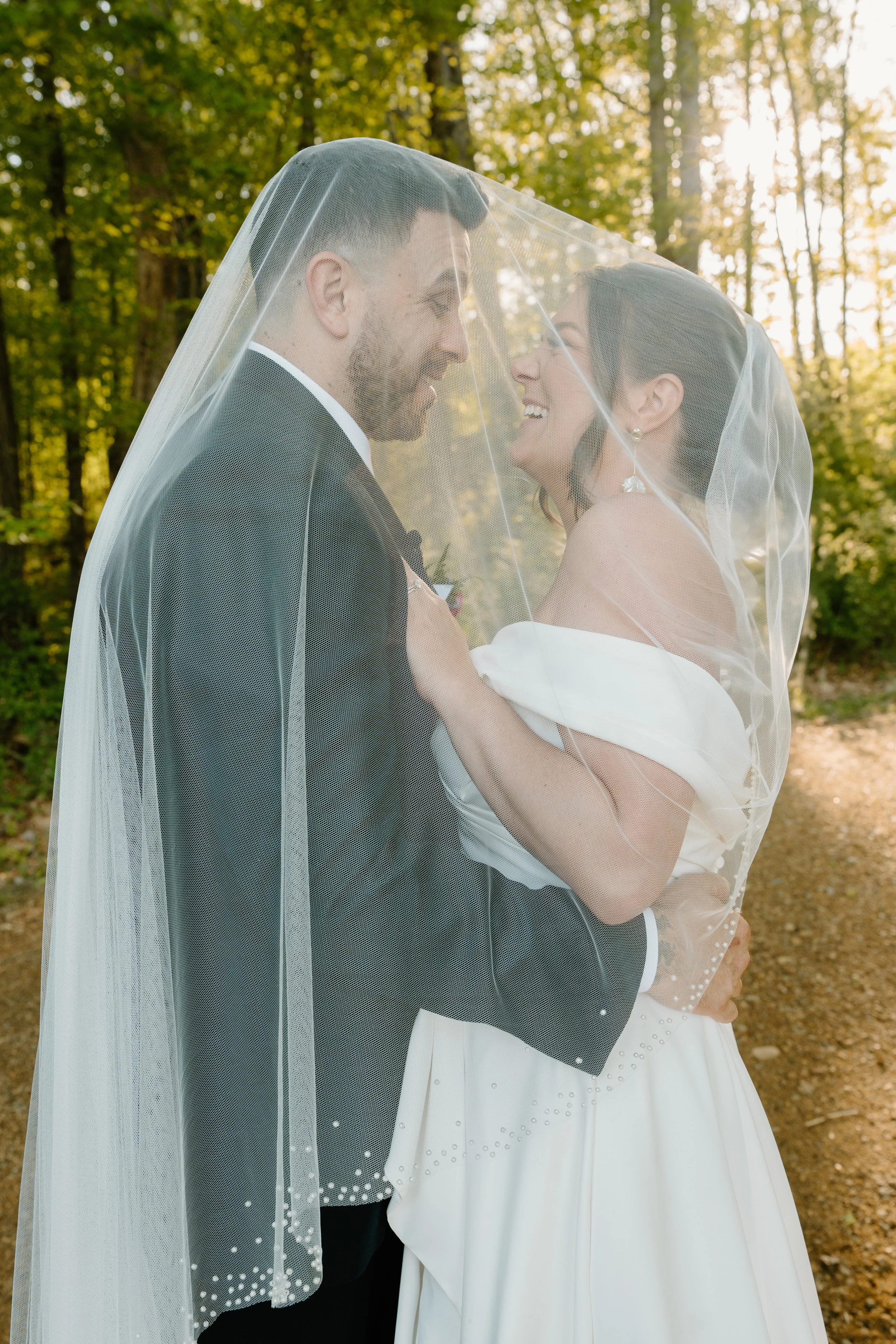 A bride and groom smiling and gazing at each other under a veil, outdoors in a wooded area for a wedding at Holiday Hill Day Camp in Connecticut.