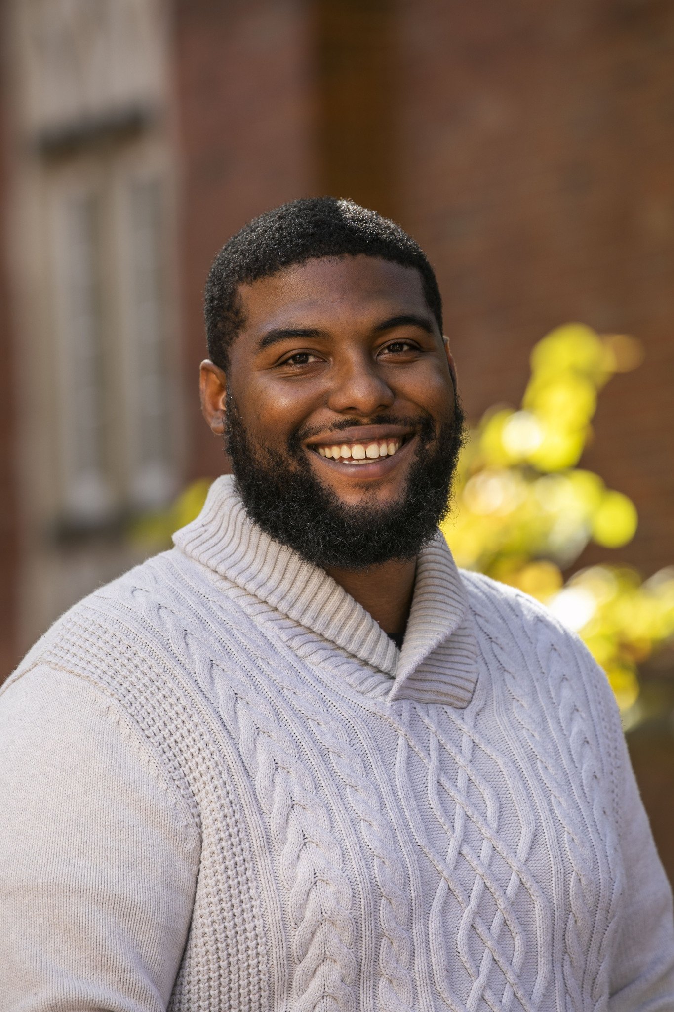 A smiling African American man with a beard and short hair, wearing a cream-colored cable-knit sweater, standing outdoors with a blurred background of trees and buildings.