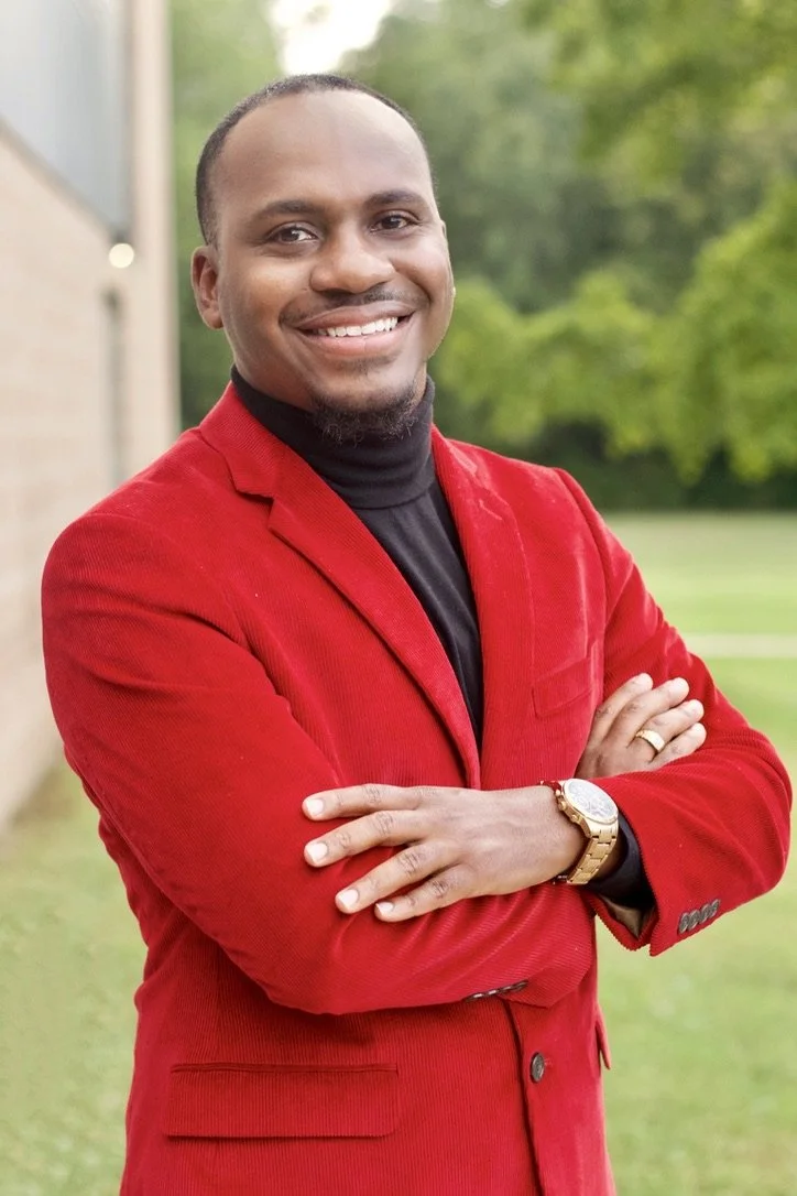 A man smiling and standing outdoors in front of trees, wearing a red blazer, black turtleneck, gold wristwatch, and pose with arms crossed.