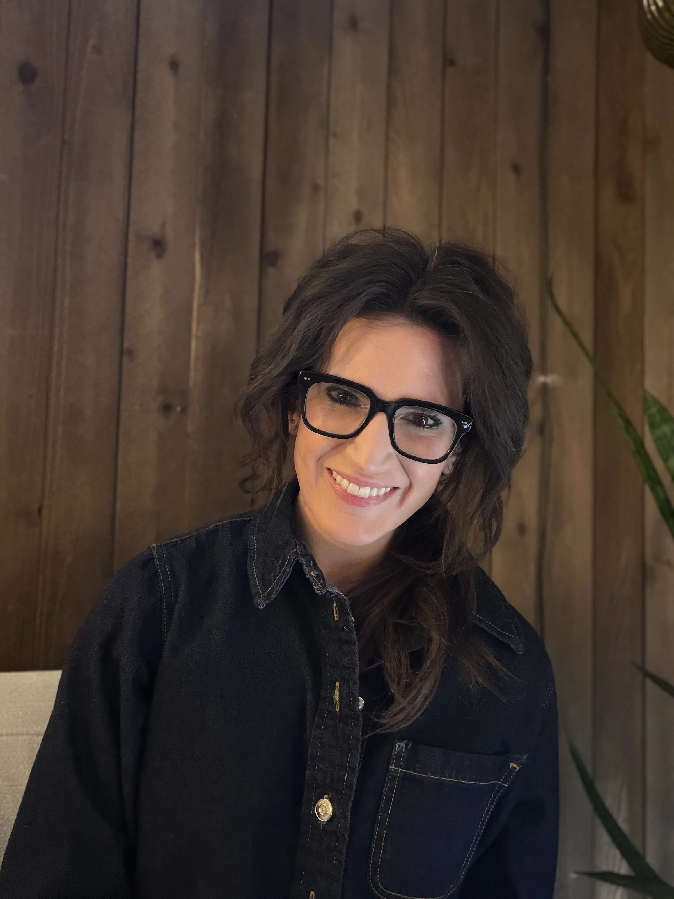 A woman with straight dark brown hair, wearing a black top and gold jewelry, smiling while sitting indoors near a white wall and potted green plants.