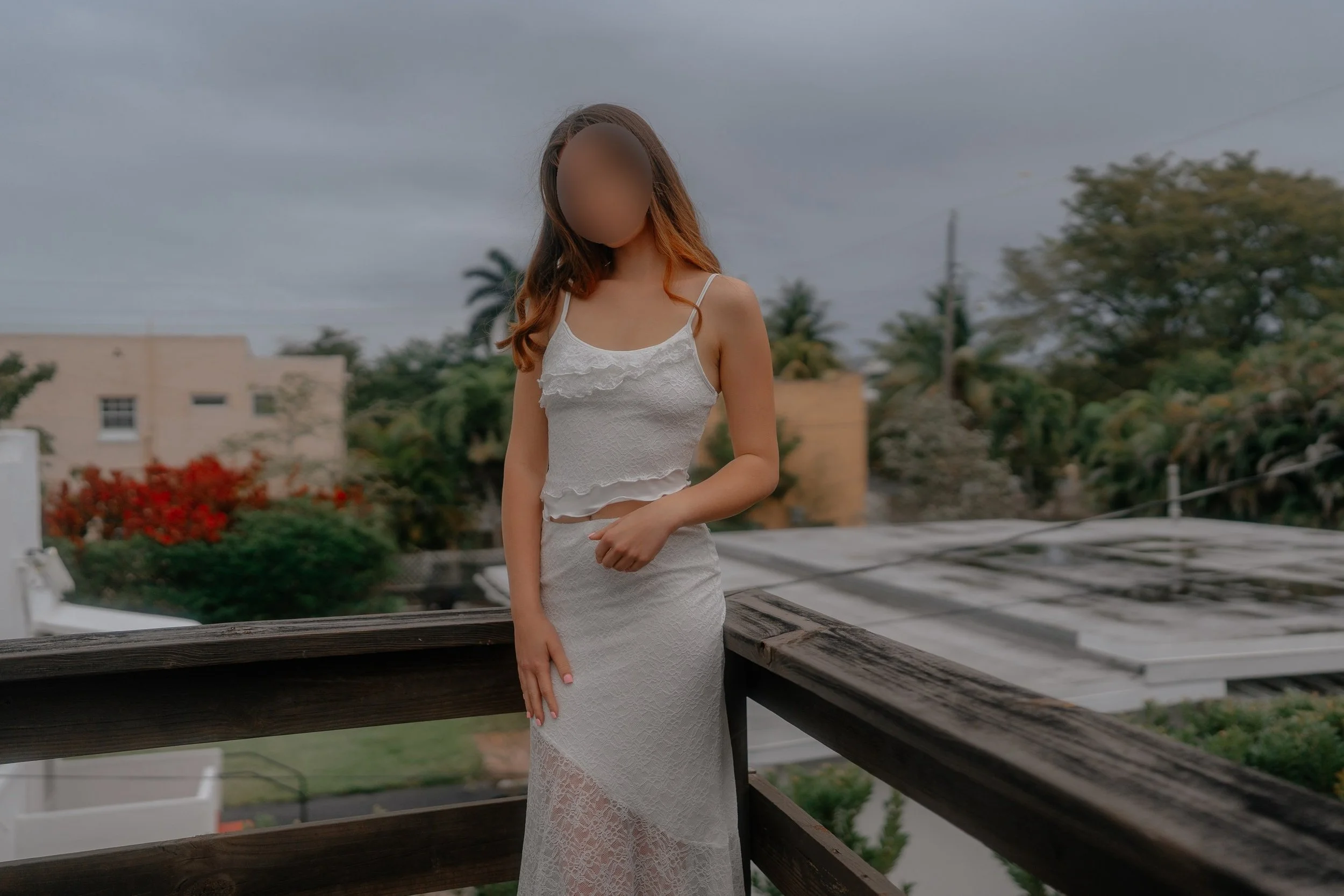 A woman in a white lace dress standing on a wooden balcony with trees and cloudy sky in the background.