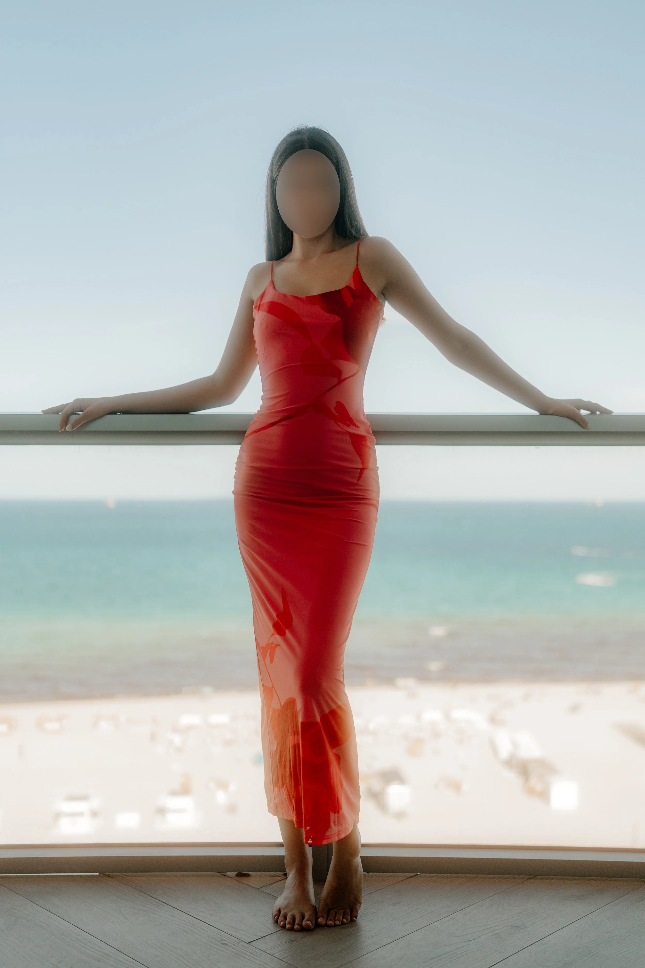 A woman in a red dress standing barefoot on a wooden floor in front of a large panoramic window overlooking the beach and ocean. The woman's face is blurred, and she is leaning slightly backward with her arms resting on the window frame.