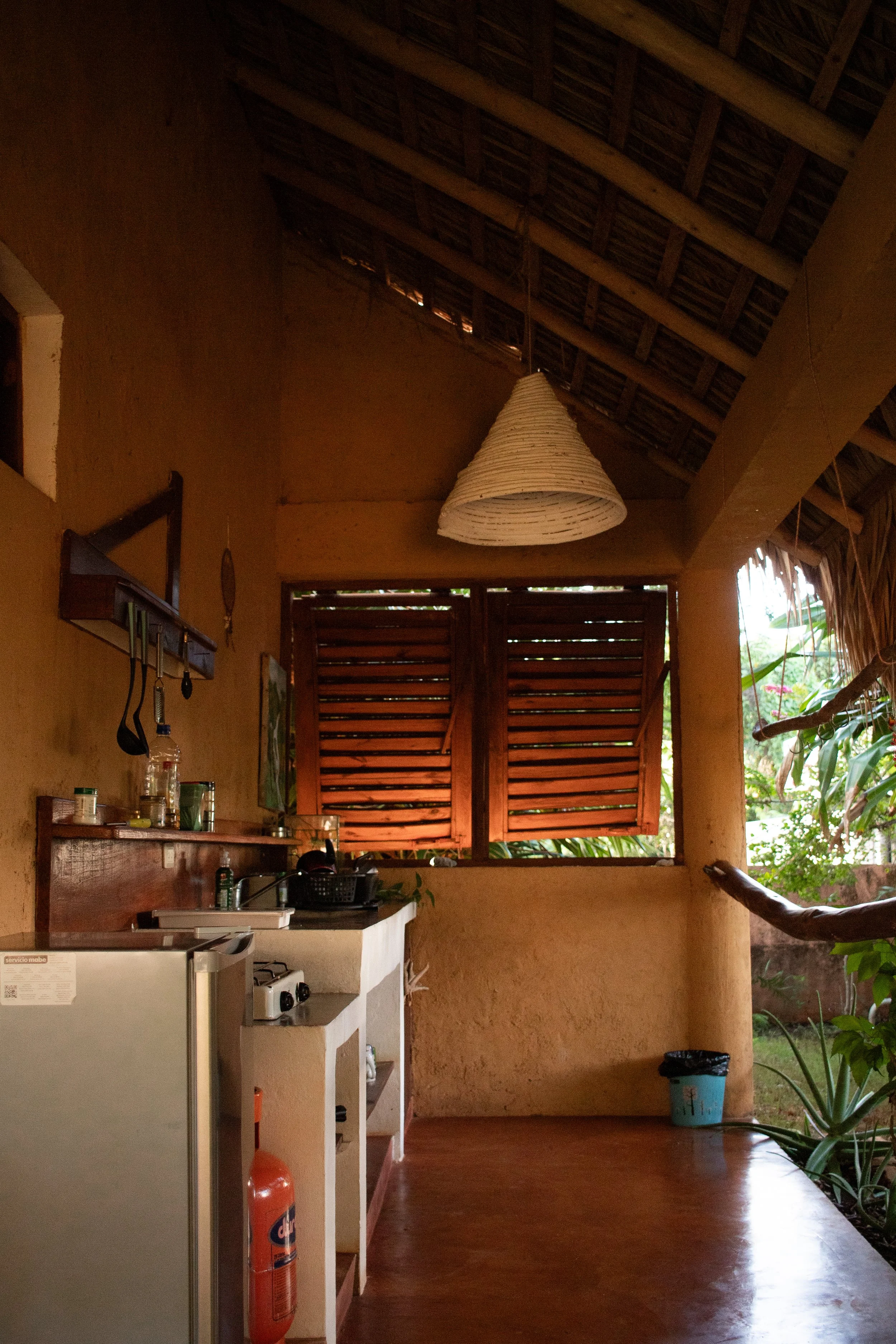 Open kitchen area with a small refrigerator, countertop, and sink. Brown wooden shutters open to outdoor greenery. Wicker lampshade hangs from a thatched roof ceiling. There is a fire extinguisher on the floor.