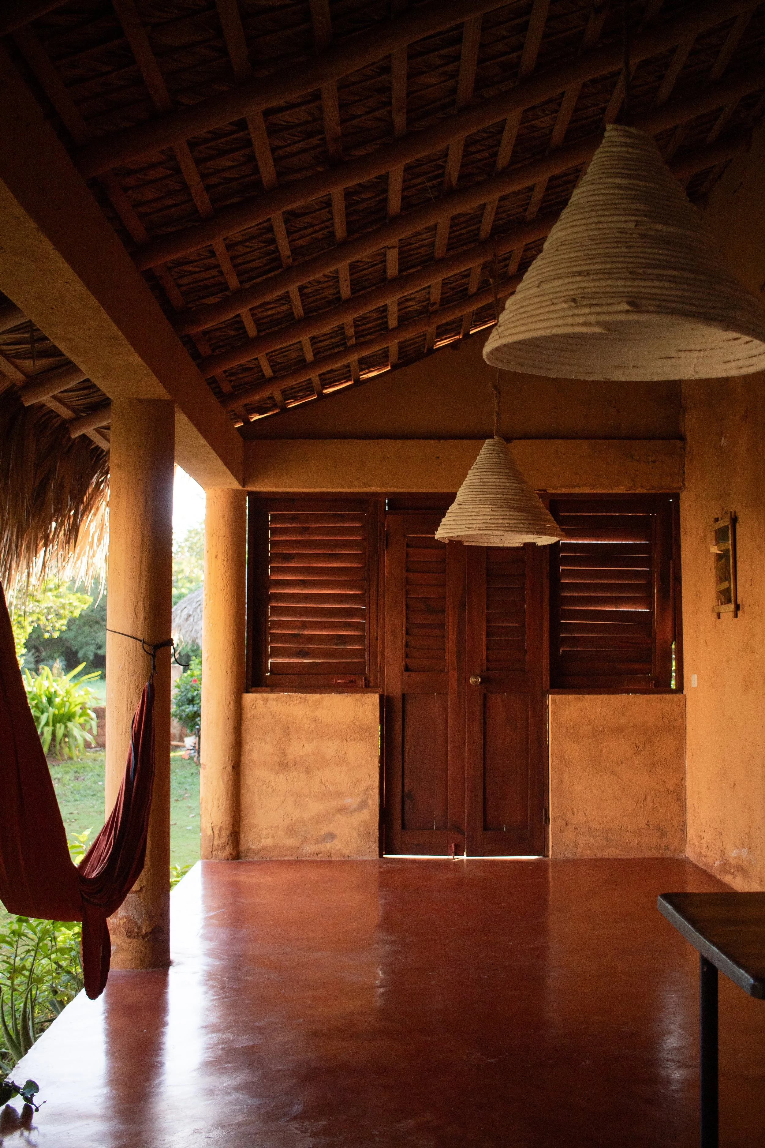 Interior of a rustic porch or room with wooden doors, shuttered windows, hanging wicker pendant lights, and a hammock on the left side.