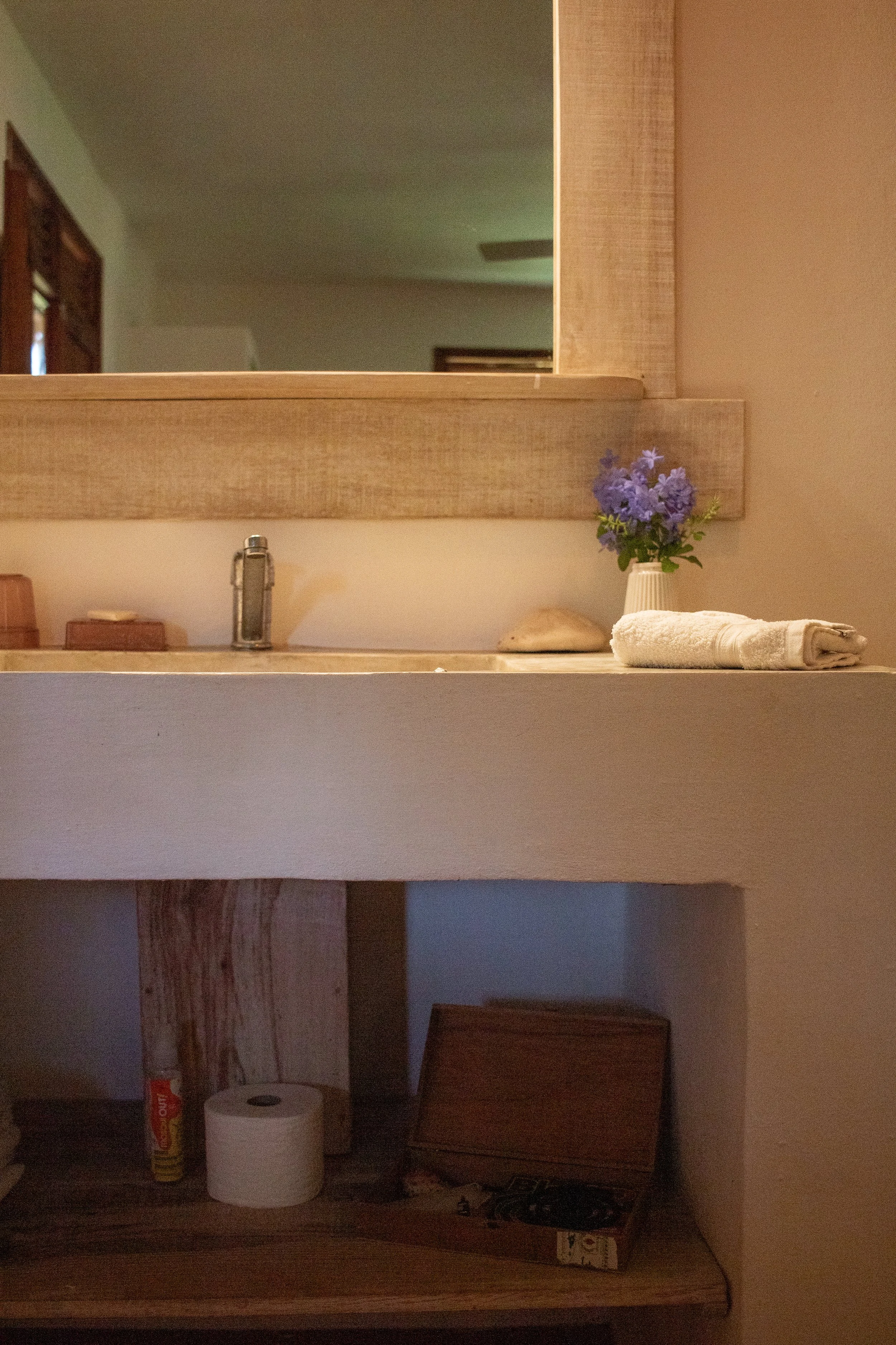 A bathroom countertop with a sink, a small vase with purple flowers, a rolled white towel, a soap dish, and some miscellaneous items underneath.