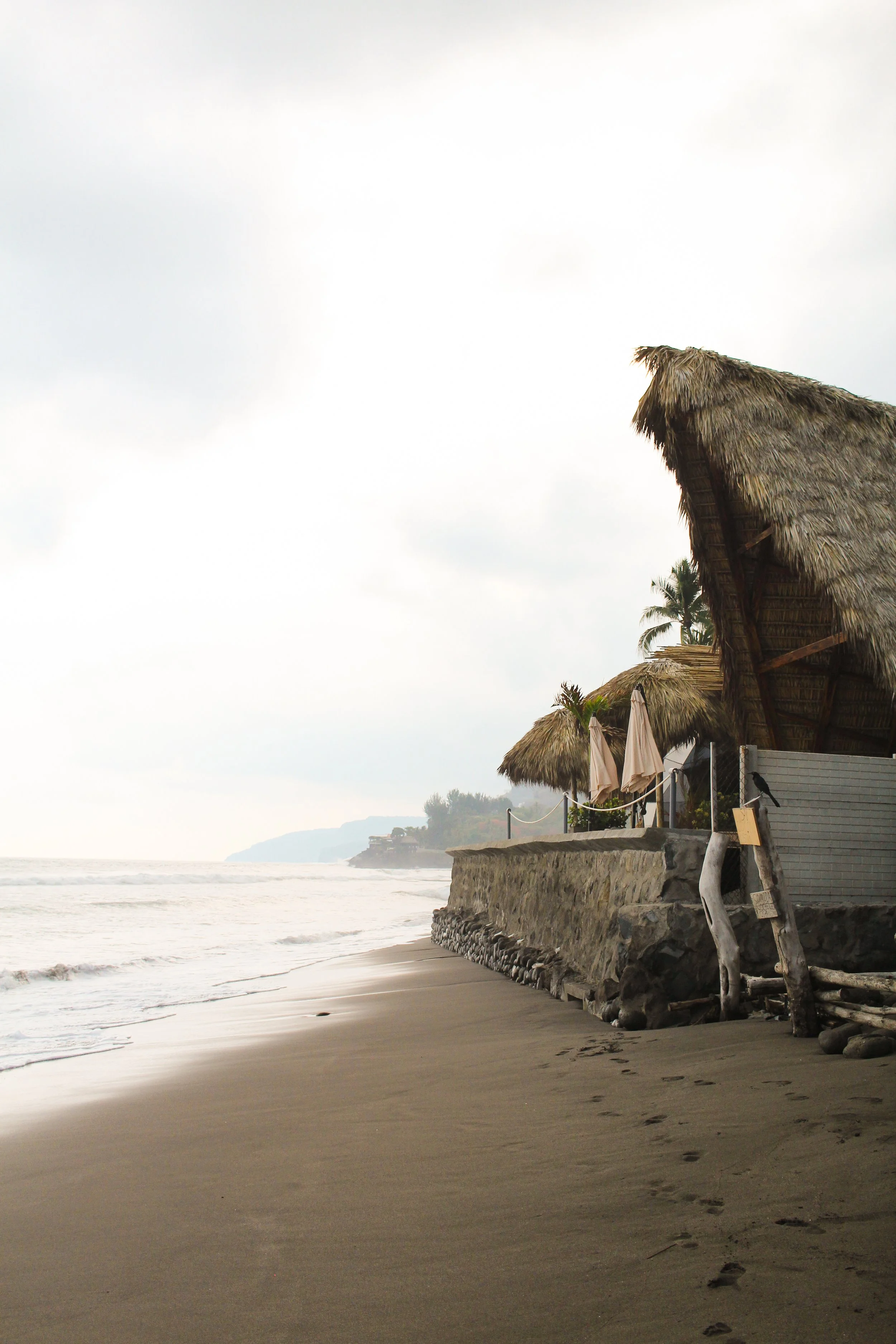 Empty beach with thatched roof tiki huts and sandy shoreline, with footprints in the sand and calm ocean waves.
