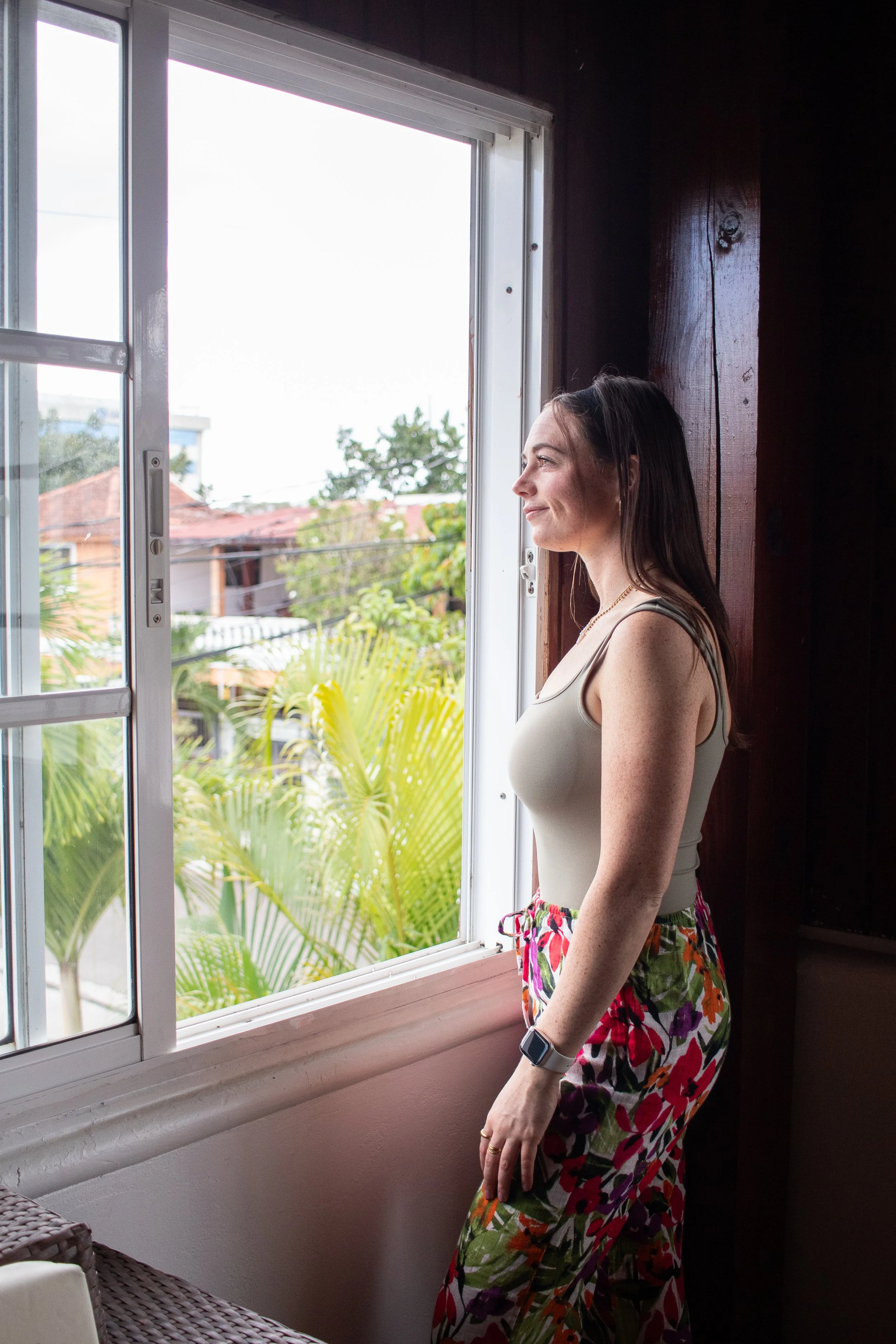 A woman stands by a window looking outside, wearing a sleeveless top, floral pants, and a smartwatch.