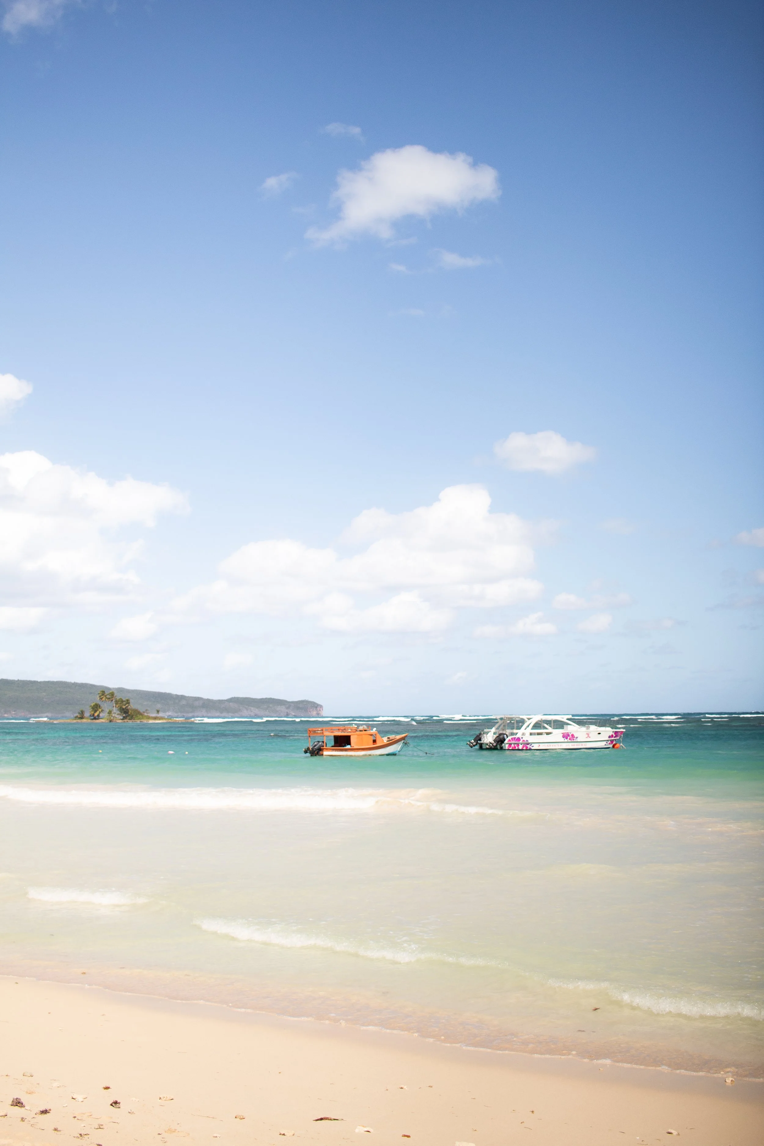 Sunny beach with white sand, turquoise water, and two boats anchored near the shore, with a small island and cloudy sky in the background.