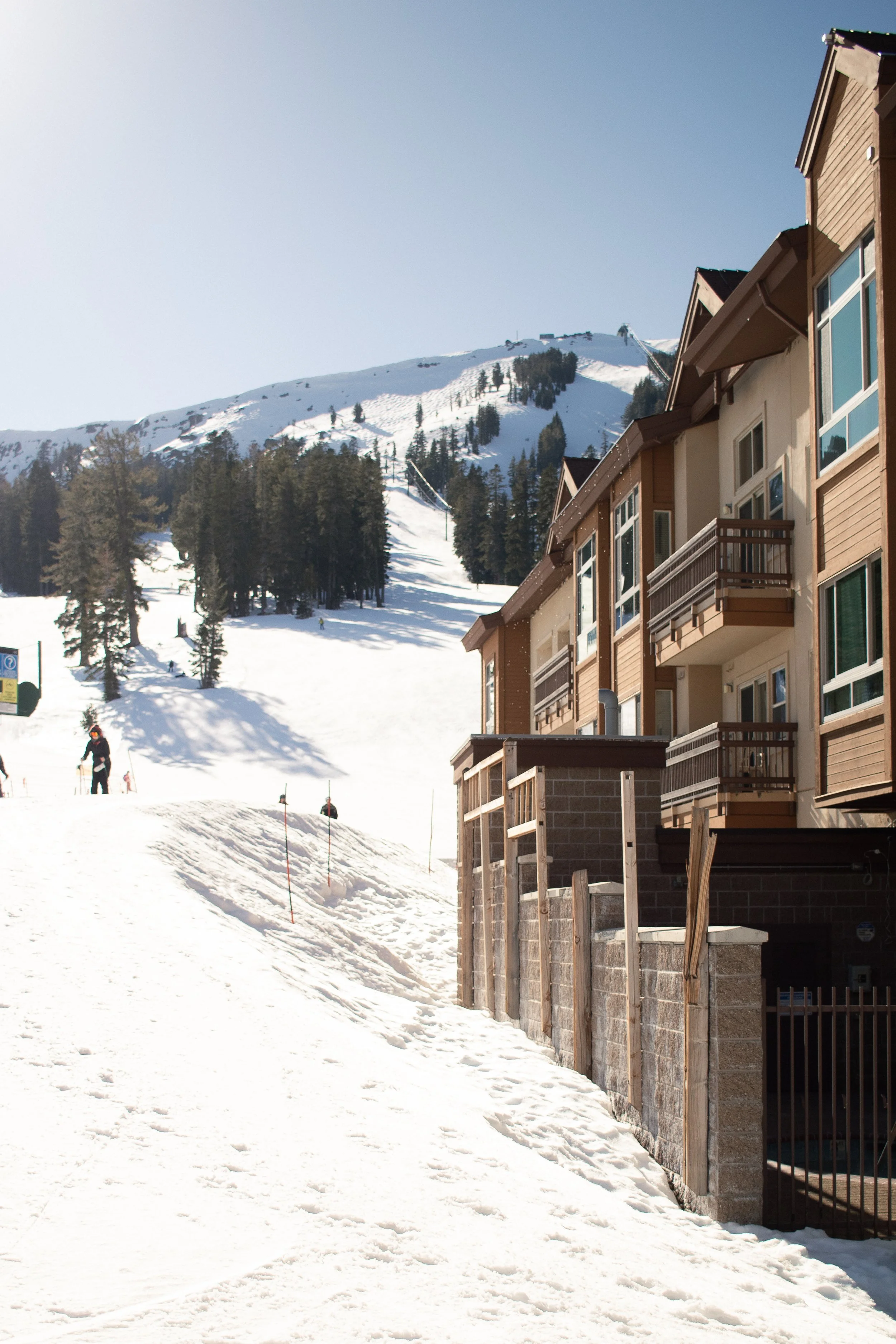 Snow-covered residential area with ski slope, skiers, and apartment buildings under clear sky in a mountain setting.