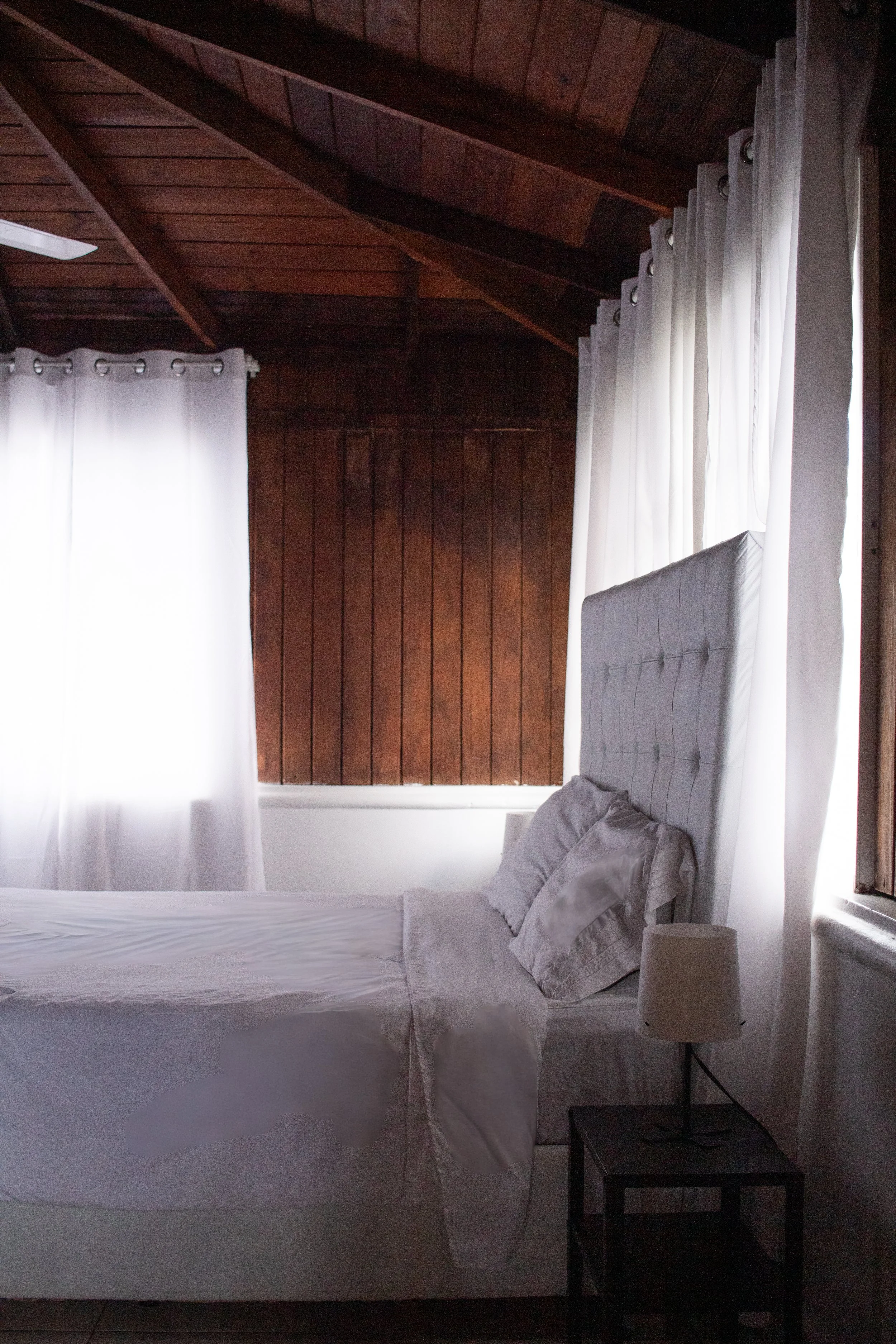 A bedroom with a white bed, pillows, and a bedside table with a lamp, featuring white curtains and a wooden ceiling and wall.