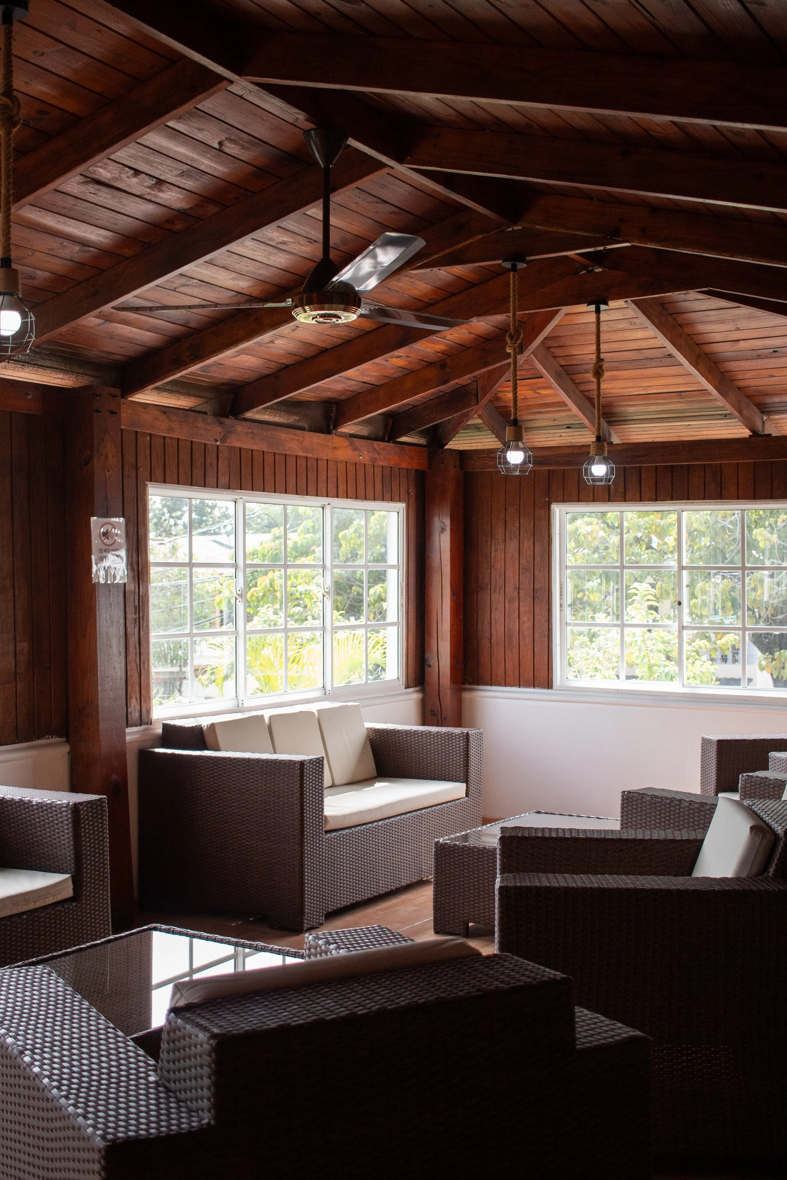 A cozy indoor seating area with wicker furniture, beige cushions, wooden walls, and a ceiling with exposed wooden beams and ceiling fan, illuminated by hanging lights near windows showing greenery outside.