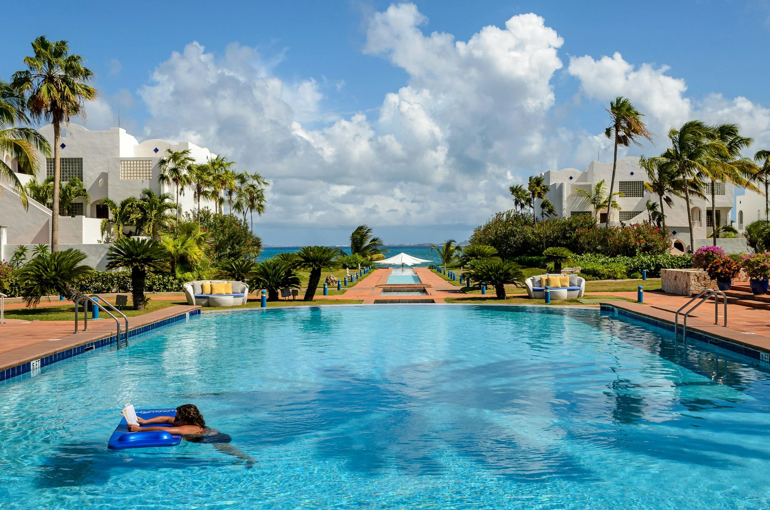 A person floating in a swimming pool with a blue float, reading a book. The pool is surrounded by tropical plants, palm trees, and white buildings. In the background, there is a view of the ocean with a blue sky and white clouds.