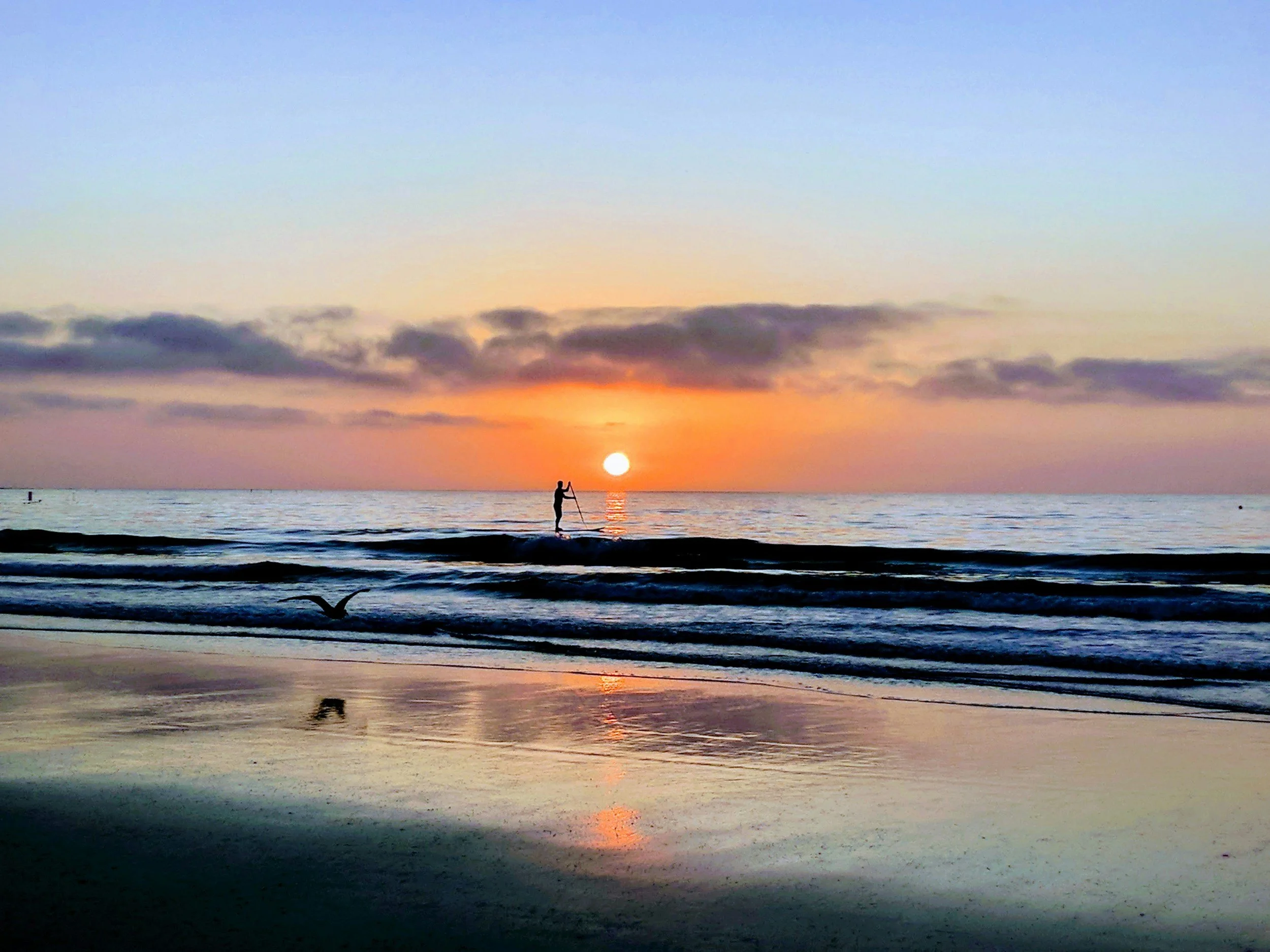 A person paddleboarding on calm ocean waters during sunset, with the sun low on the horizon, colorful sky, and a few clouds.