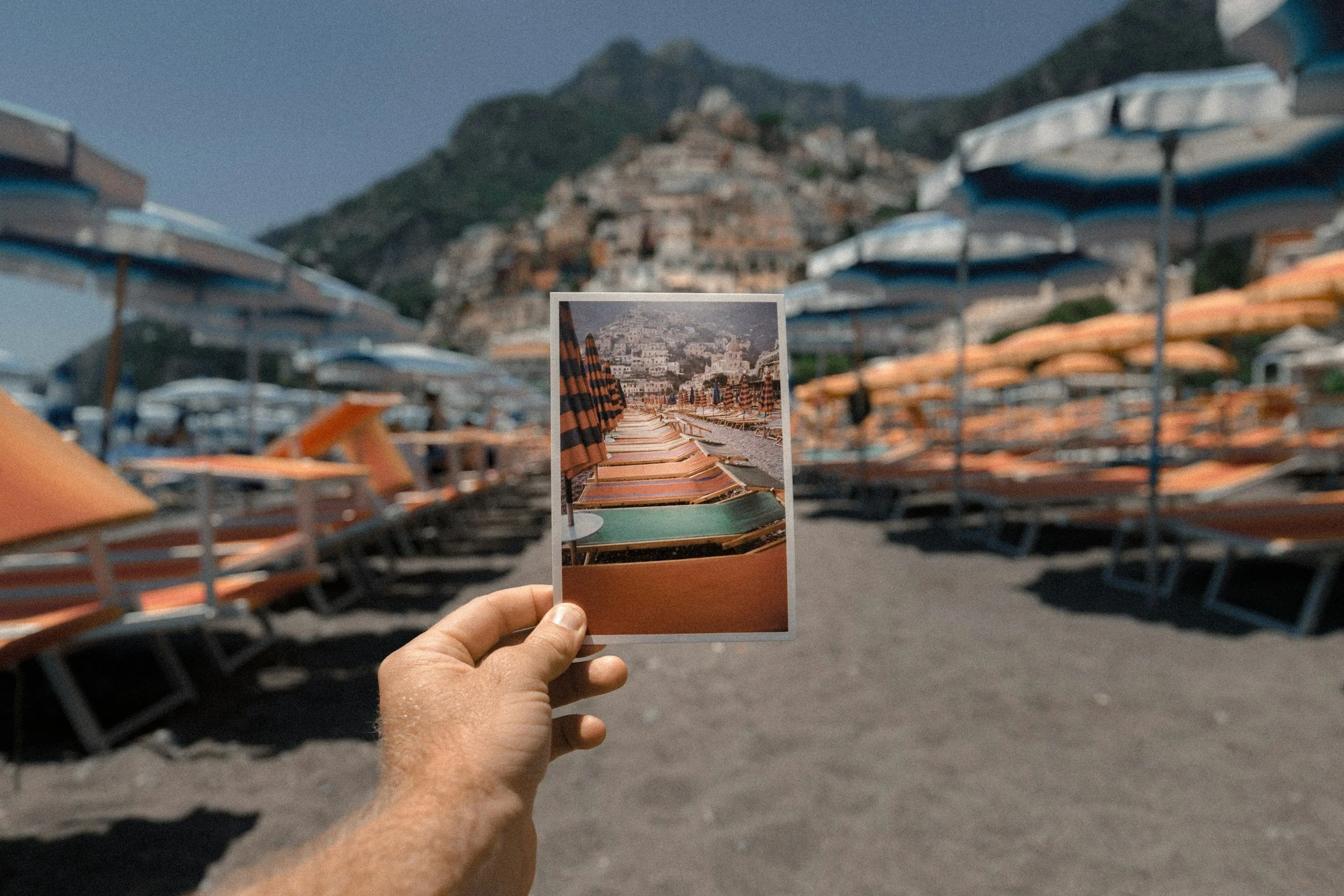 A person holding a photograph of a beach scene with striped umbrellas and lounge chairs, with rows of similar umbrellas on the actual beach in the background and a mountain in the distance.