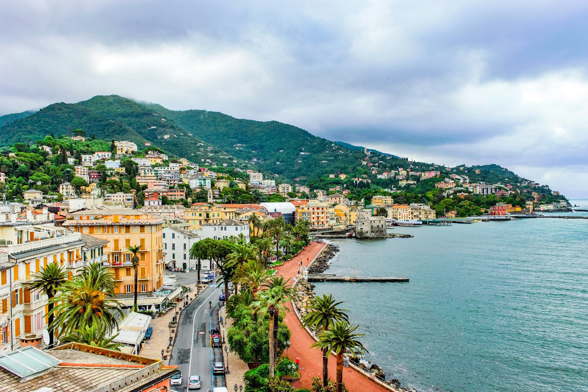 A scenic coastal city with colorful buildings along the shoreline, a road with parked cars and palm trees, and lush green hills in the background under a cloudy sky.