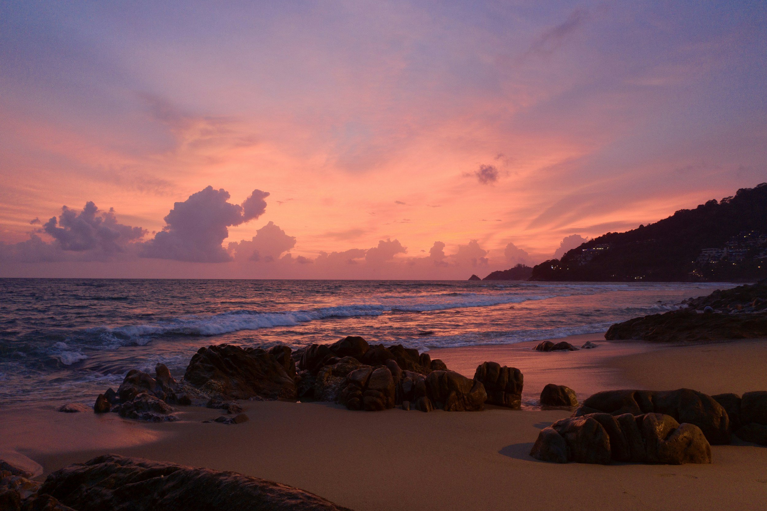 Sunset over the ocean with a colorful sky of pink and purple hues, clouds, and a coastline with rocks and buildings on a hillside in the distance.