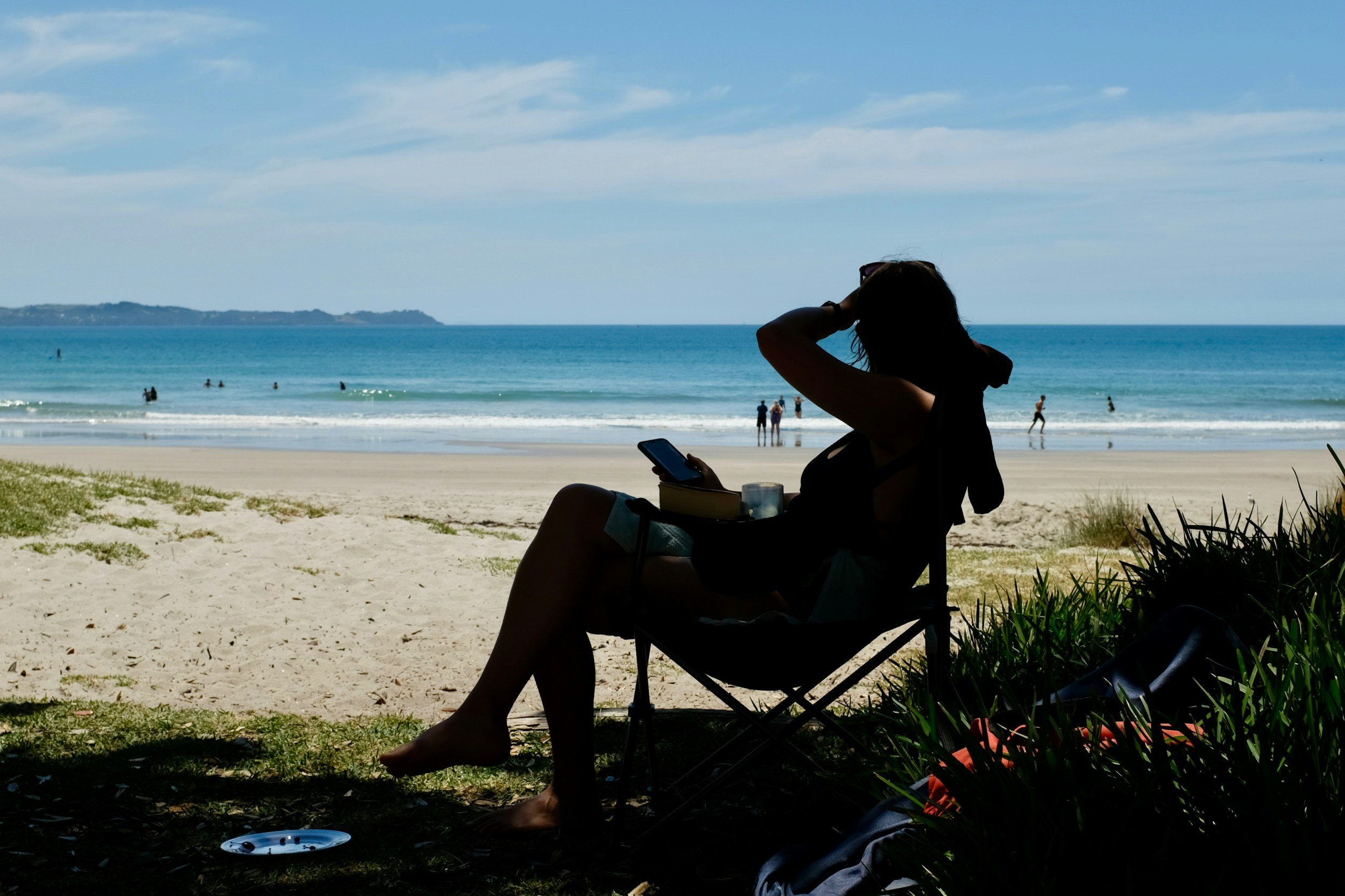 A woman sitting on a beach in the shade, looking at her phone, with the ocean and people swimming in the background.