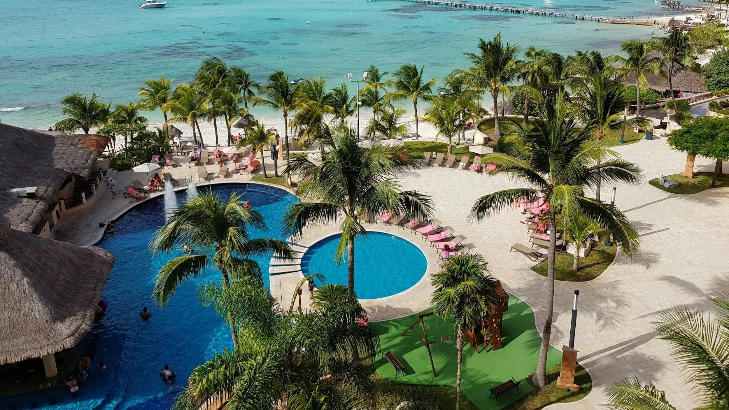 A view of a tropical resort with two swimming pools, palm trees, lounge chairs, and umbrellas near a beach with turquoise water.